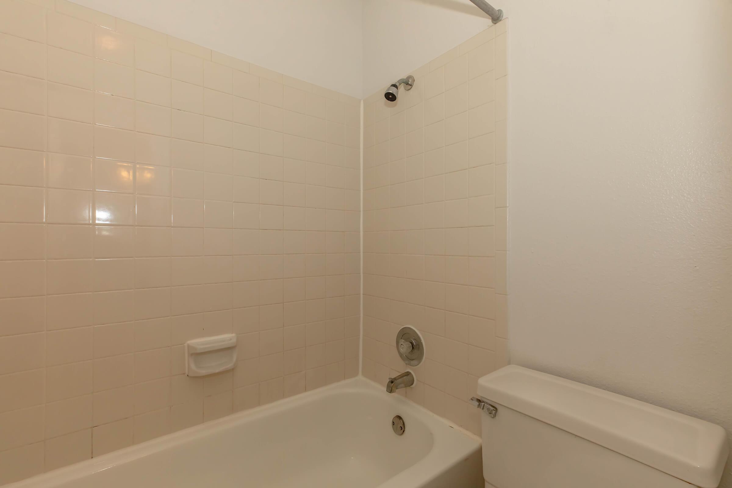 A simple bathroom featuring a white bathtub with a showerhead above it, beige tiled walls, and a small storage shelf. A toilet is positioned next to the bathtub, all set against a plain white background. The overall appearance is clean and functional.