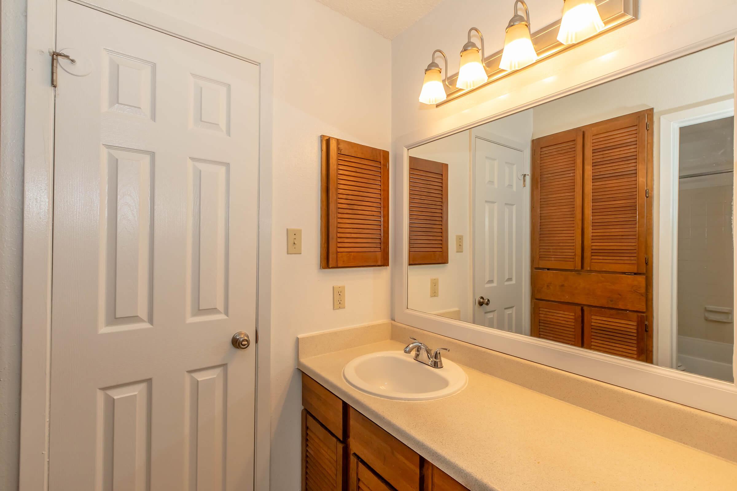 A small bathroom featuring a sink with a countertop, a large mirror above the sink, and wooden cabinets with slatted doors. Light fixtures above the mirror provide illumination. A white door is visible on the left side, and a light-colored wall completes the simple design.