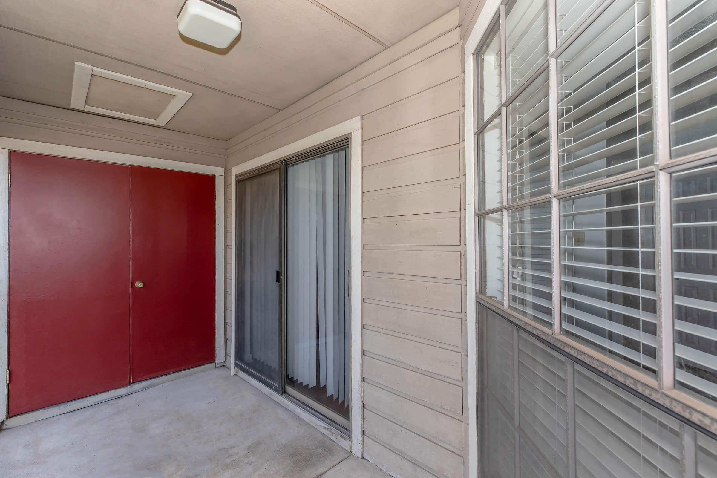 A view of an apartment entrance featuring two red doors, a sliding glass door with sheer curtains, and a window with wooden blinds. The walls are textured and painted in a light color, and the ceiling is equipped with a light fixture. The floor is concrete, creating a simple, modern entryway.