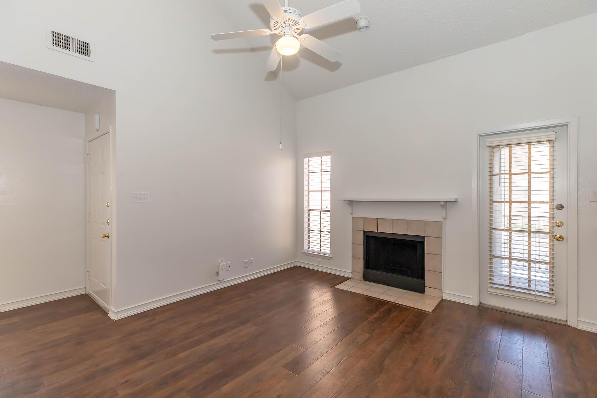 Spacious living room featuring a tall ceiling with a ceiling fan, light-colored walls, and hardwood flooring. A fireplace is situated in the corner, and large windows with blinds allow natural light to enter the room. There are two doors leading to an outside patio.