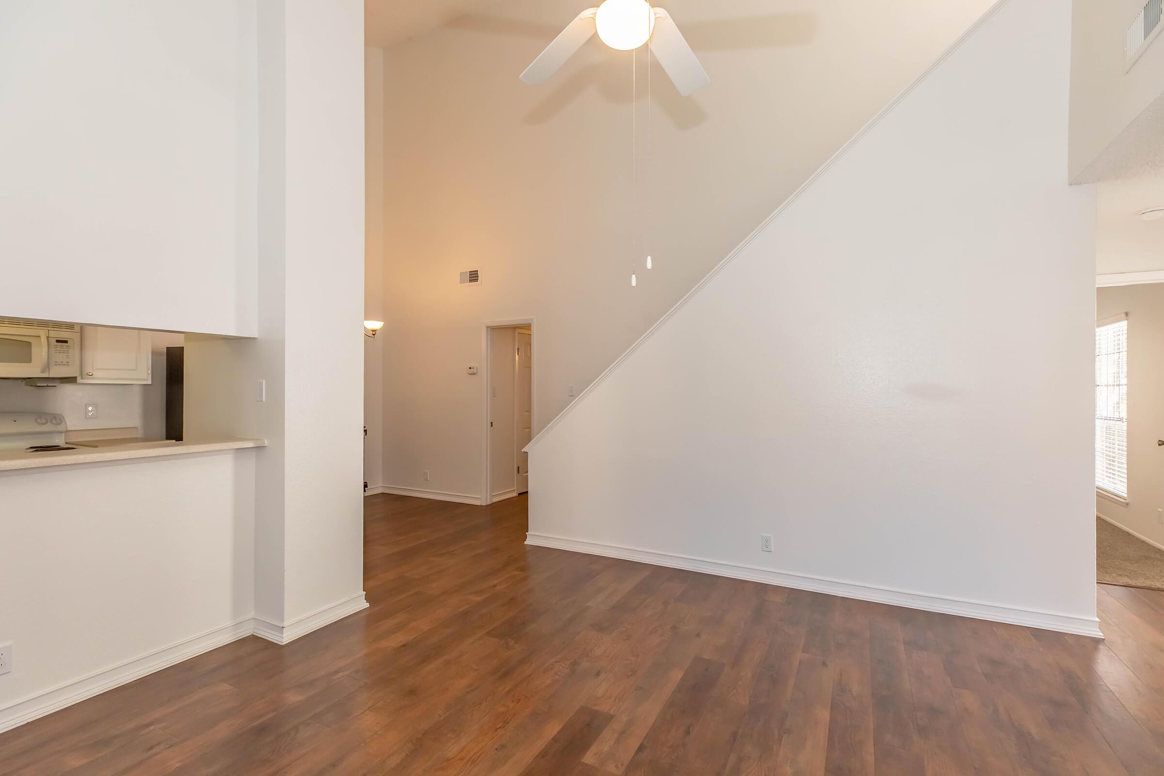 A spacious interior of a modern living area featuring high ceilings, a ceiling fan, and hardwood flooring. To the left, a kitchen area is visible with a bar counter, and a staircase leads to an upper level. Natural light enters through windows, contributing to a bright and airy atmosphere.