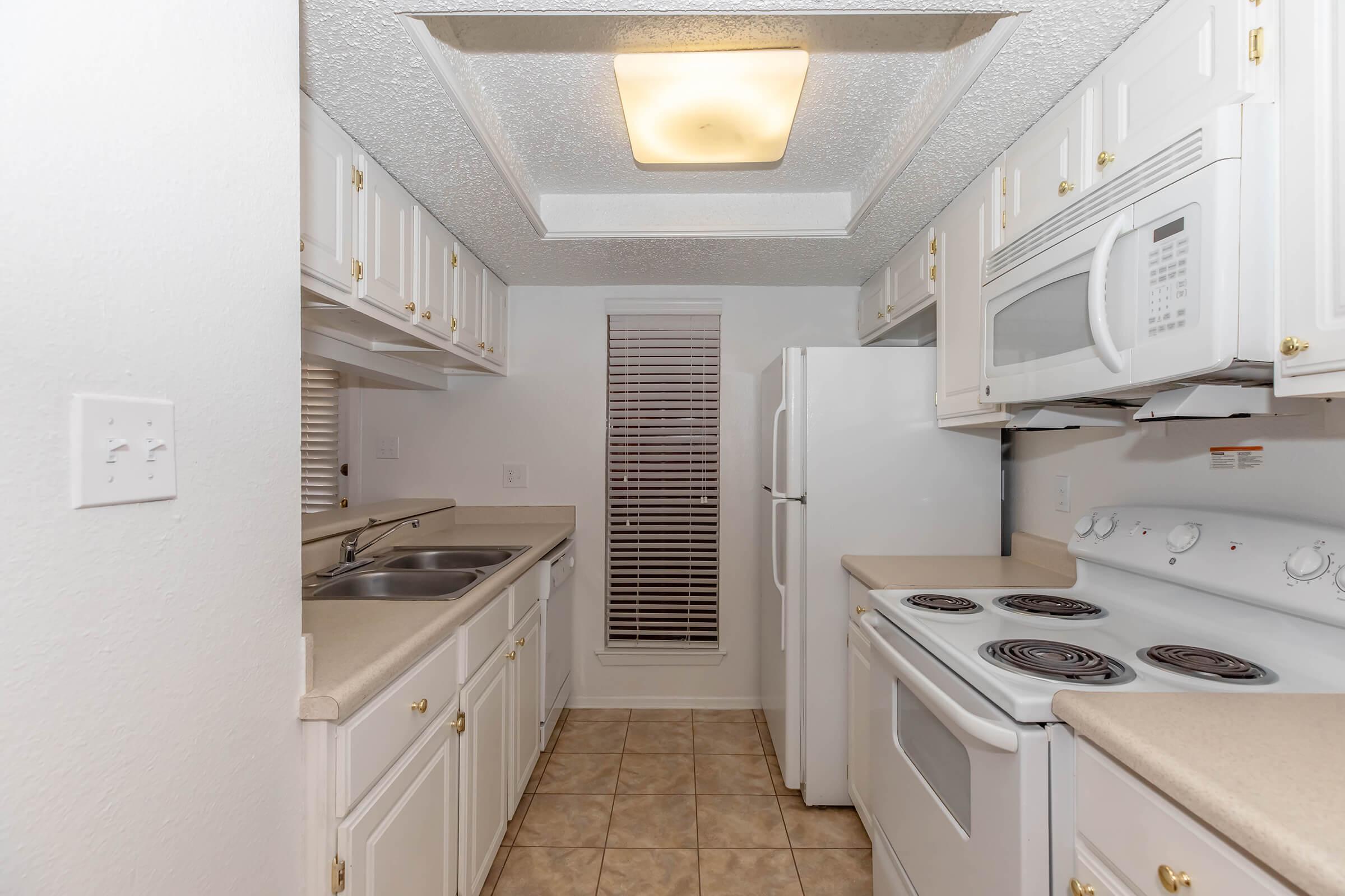 A small, well-lit kitchen featuring white cabinetry with gold handles, a double sink, a refrigerator, a microwave, and an electric stove. The countertops are beige, and there is a window with blinds providing natural light. The floor is tiled in a light color, complementing the overall clean and simple design.