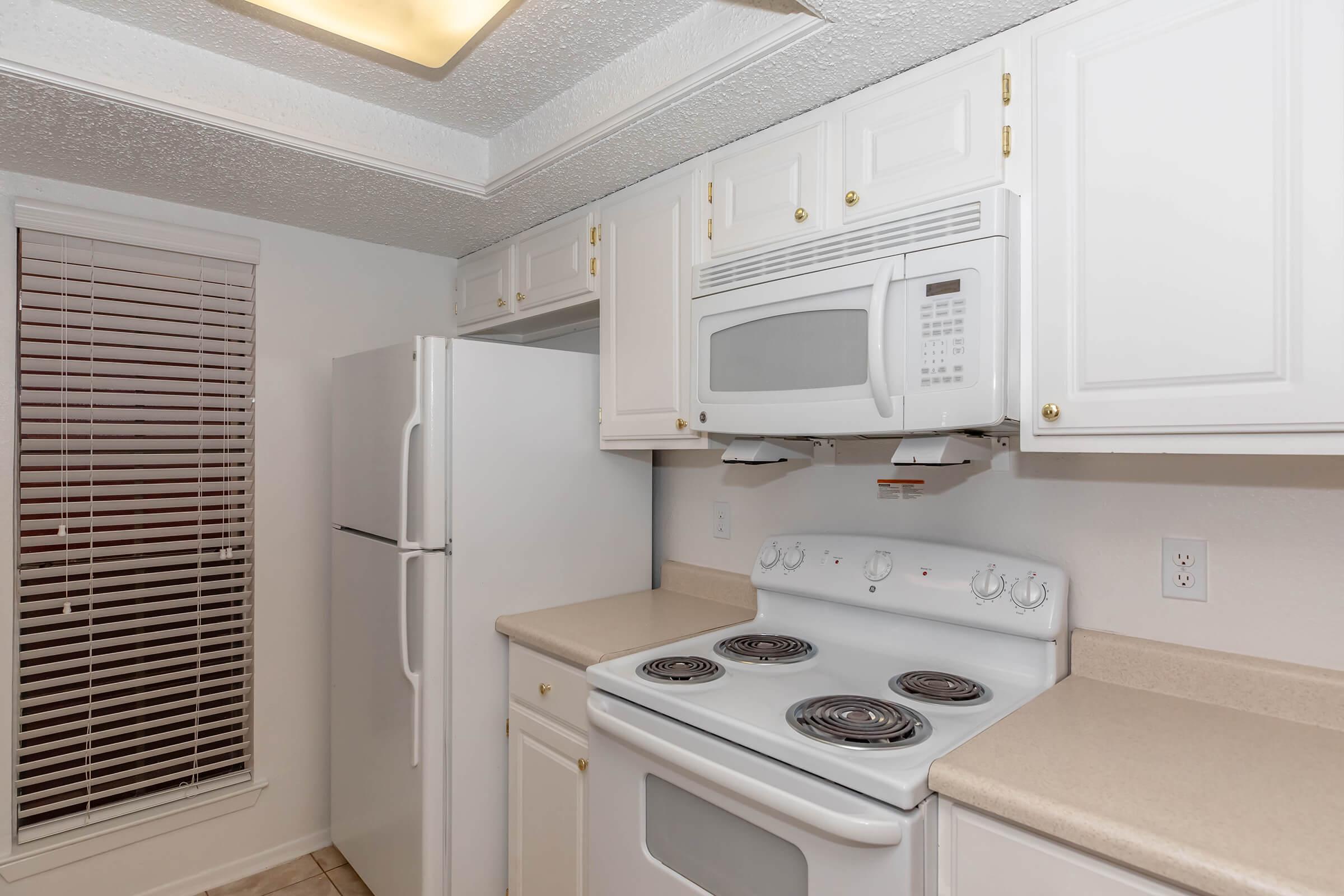 A kitchen featuring white cabinets, a white refrigerator, a white microwave above a white stove with coil burners. There is a light fixture on the ceiling and a window with blinds. The countertops are light-colored, and the overall aesthetic is clean and modern.