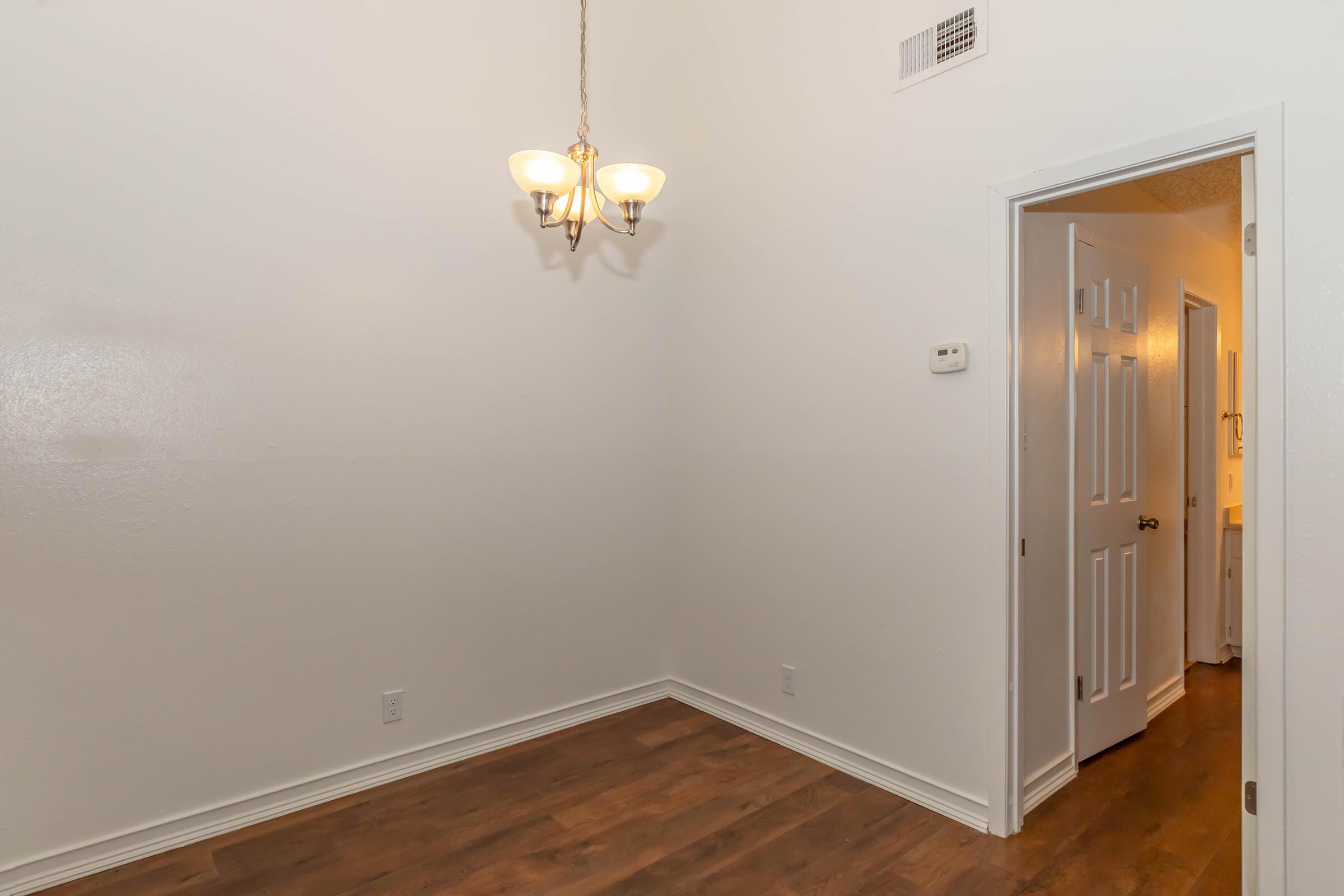 A room with light-colored walls and a wooden floor. A chandelier hangs from the ceiling, providing lighting. There is an open door leading to another room, possibly a bathroom, visible on the right side of the image. The overall atmosphere is bright and spacious.
