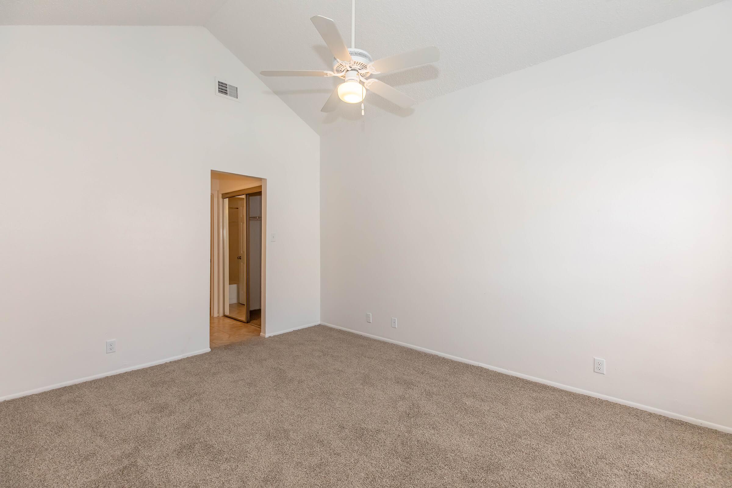 A spacious, empty room with beige carpet and white walls. The ceiling is high with a ceiling fan installed. A doorway on the left leads to a bathroom, while the overall atmosphere is bright and minimalist.