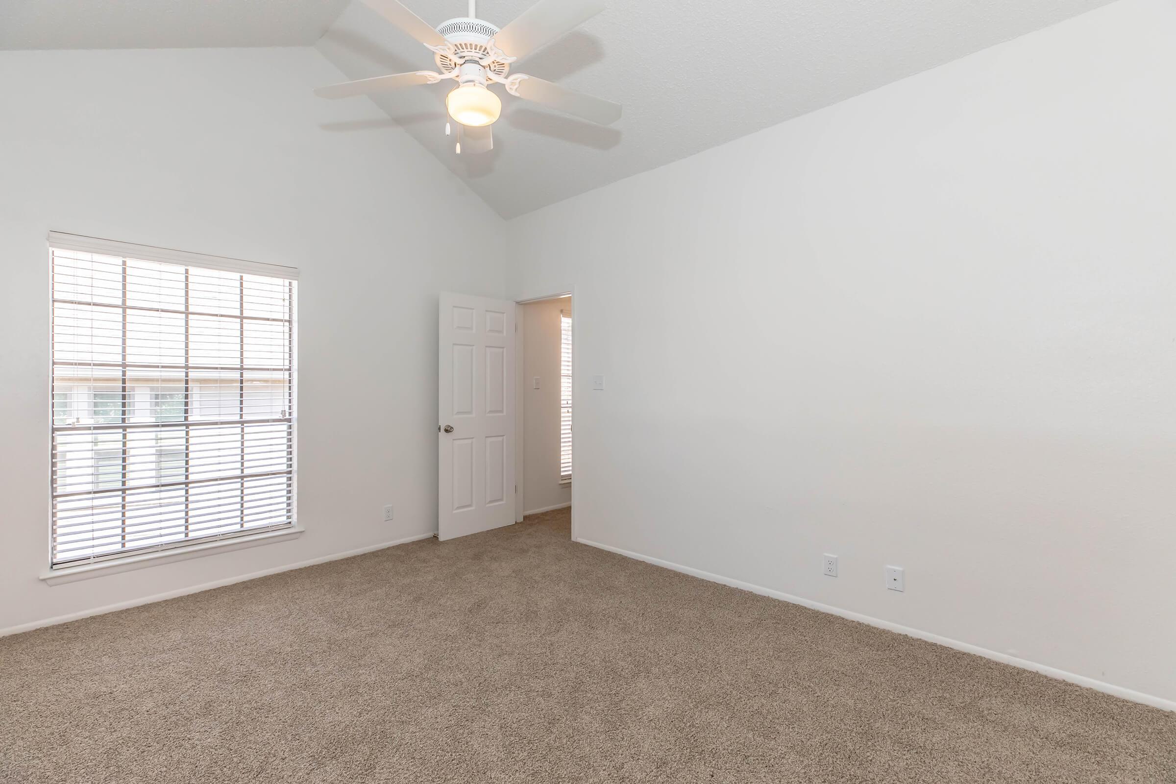 A well-lit room with a high ceiling and a ceiling fan. The walls are painted white, and the floor is carpeted in a light beige color. There are two windows with blinds on the left side, and a doorway leads to another room on the right. The overall atmosphere is spacious and airy.