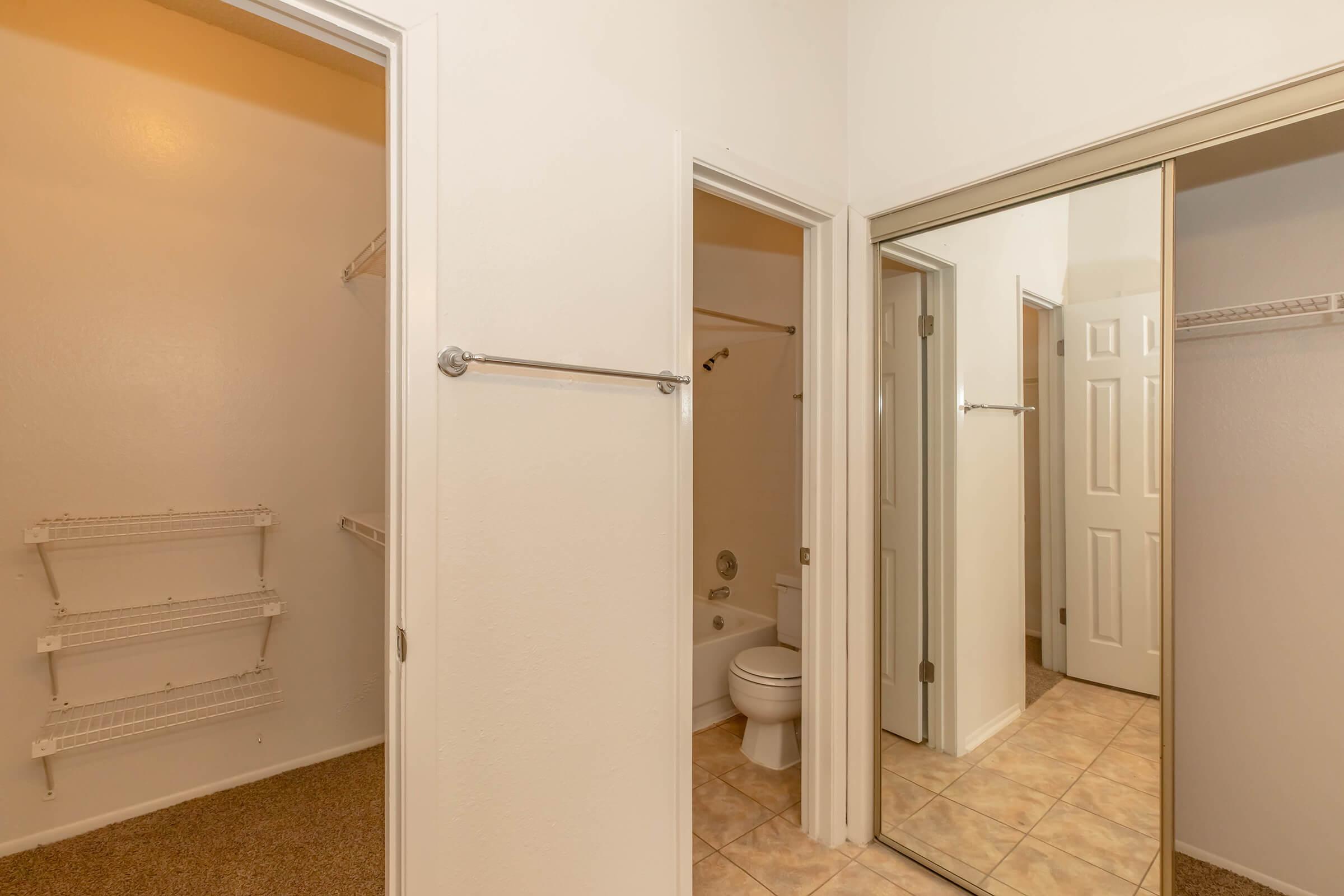 A view of a bathroom and a walk-in closet area. The bathroom features a toilet and a shower/bathtub combination, while the closet has shelving on the wall. The interior has neutral-colored walls and tiled flooring. There are mirror doors reflecting the space.