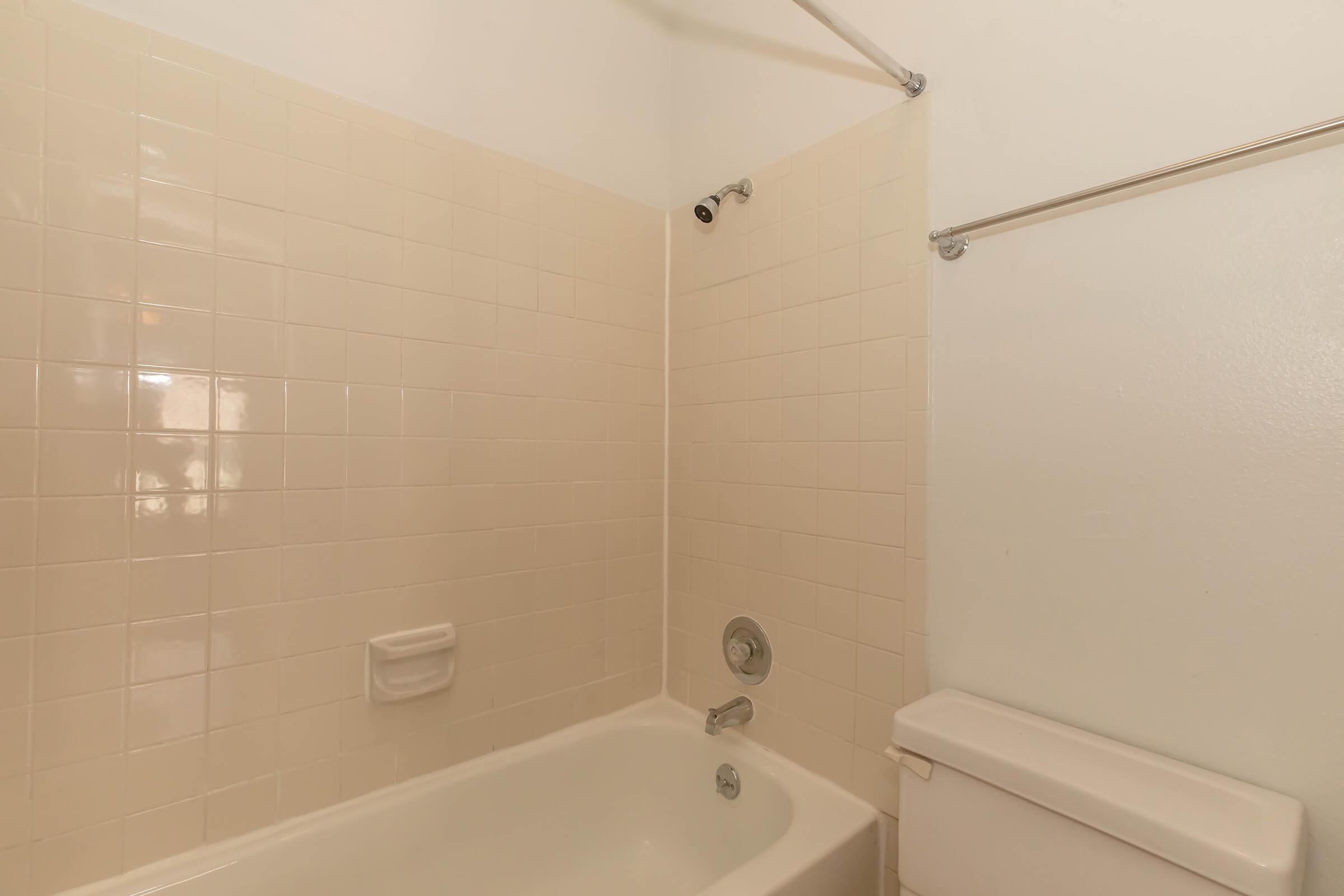 A simple bathroom with a beige tiled shower and bathtub. The tub is empty, and there's a chrome showerhead installed. The wall features a towel rack and a soap dish. The overall color scheme is light and neutral, creating a clean and minimalist atmosphere.