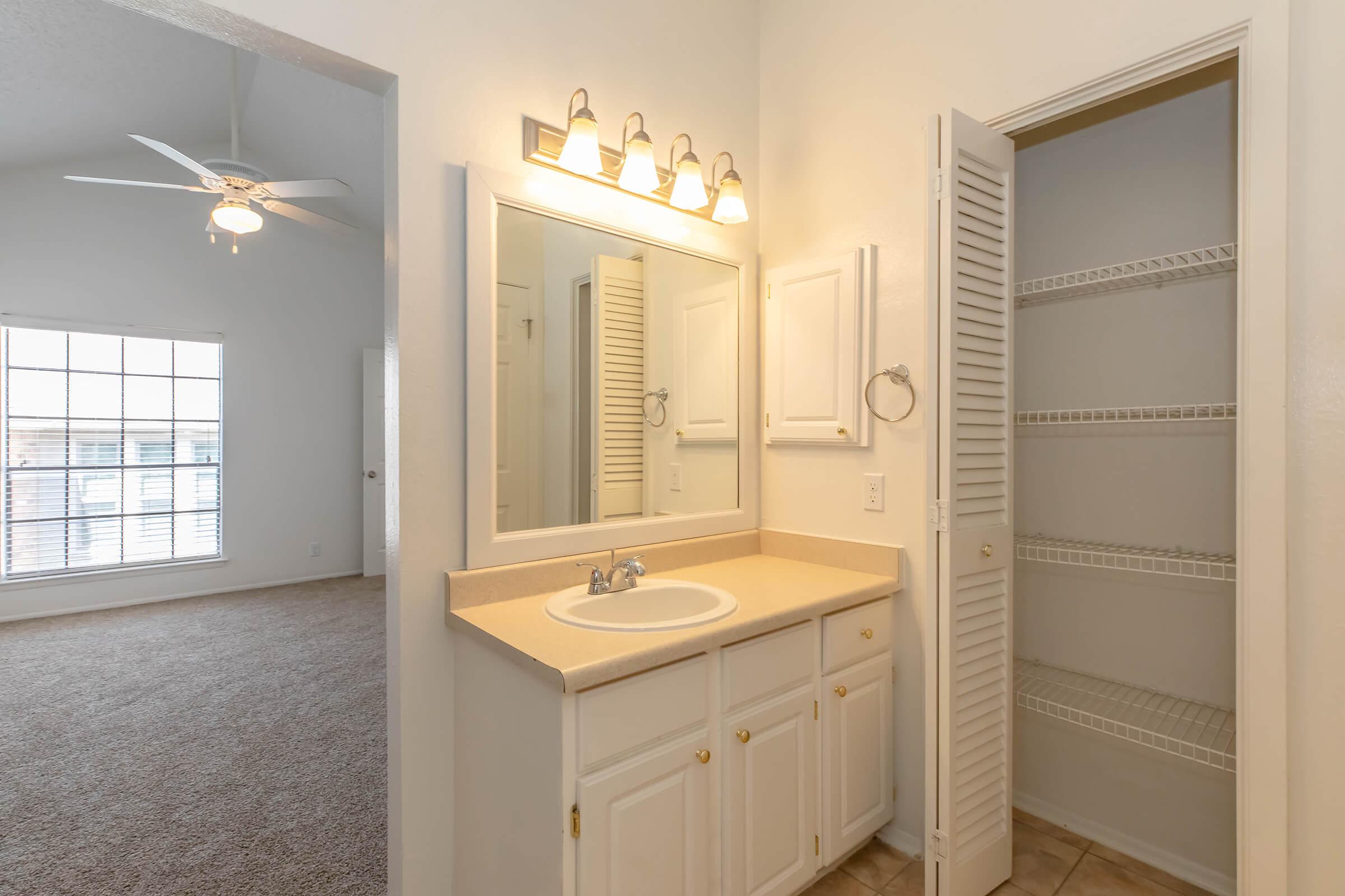 A well-lit bathroom with a cream-colored countertop, sink, and mirror. There are white cabinets below the sink, a towel ring on the wall, and a louvered closet door, revealing shelves inside. The background shows the entrance to a spacious living area with large windows and carpeted flooring.