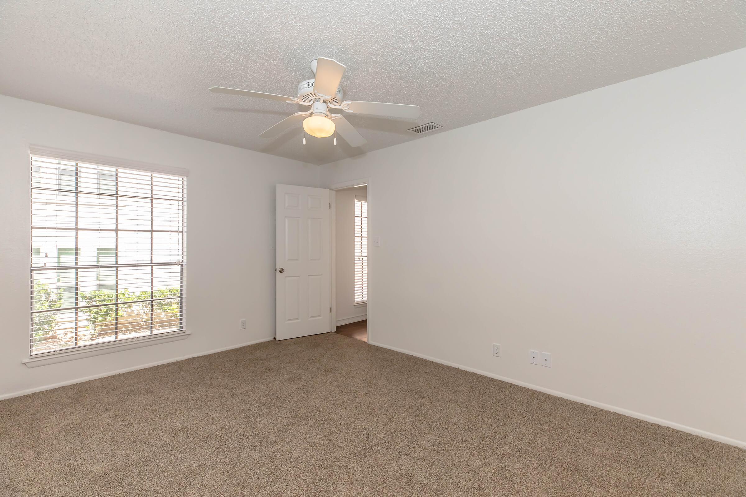 A spacious, well-lit room with beige carpet and white walls. Features include a ceiling fan, a window with blinds allowing natural light, and a door leading to another area. The room is empty, suggesting potential for personalization and decoration.