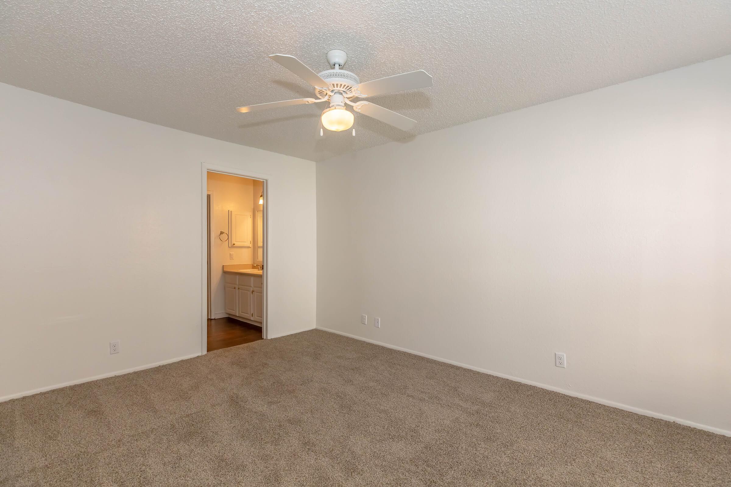 Empty room with light gray carpet, a ceiling fan, and white walls. A doorway leads to another room, which appears to have a bathroom with light-colored cabinetry. Natural light illuminates the space, creating a bright and open atmosphere.