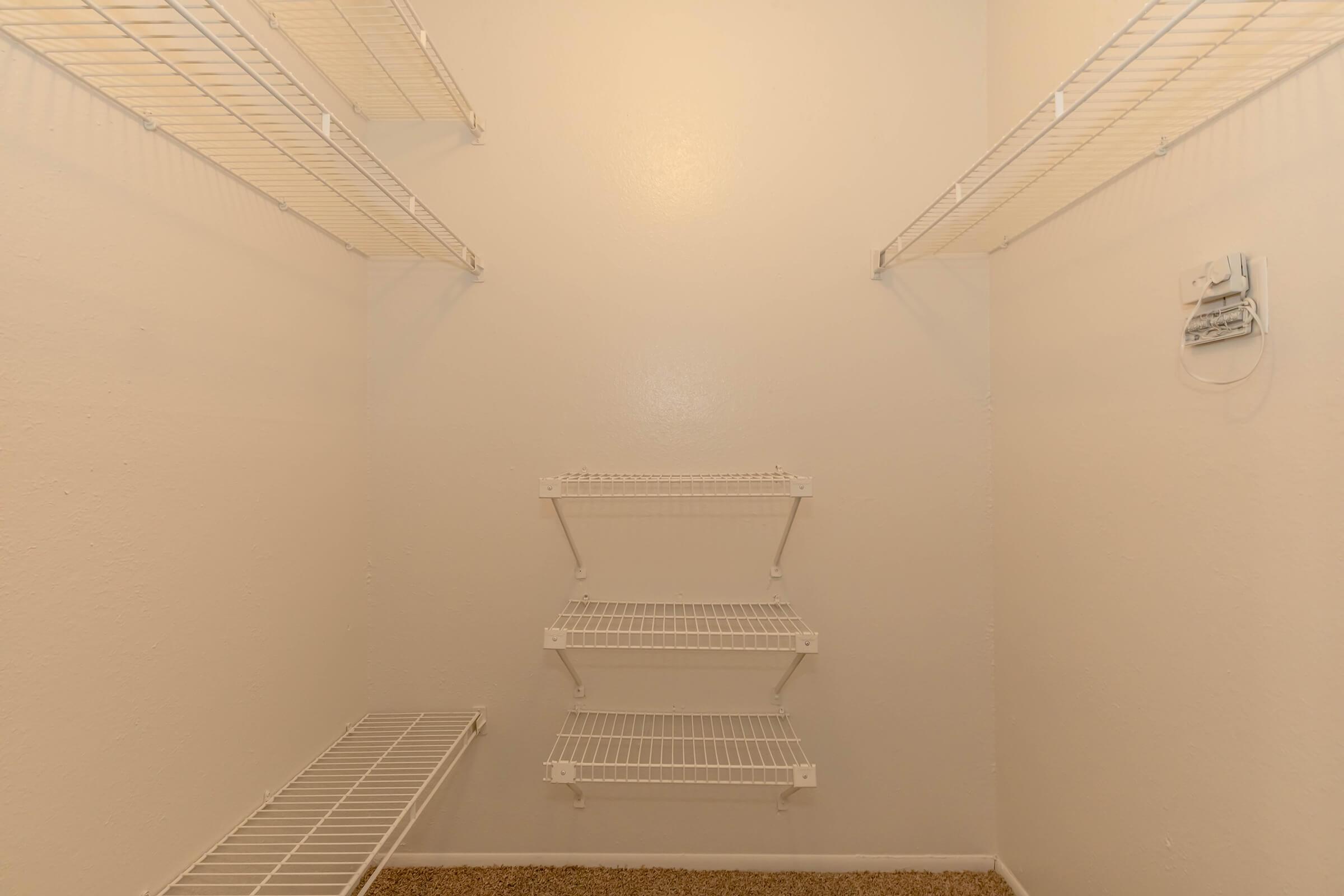 Empty walk-in closet featuring white wire shelving mounted on the walls. The closet has a light-colored wall and carpeted flooring, with several shelves arranged for storage.
