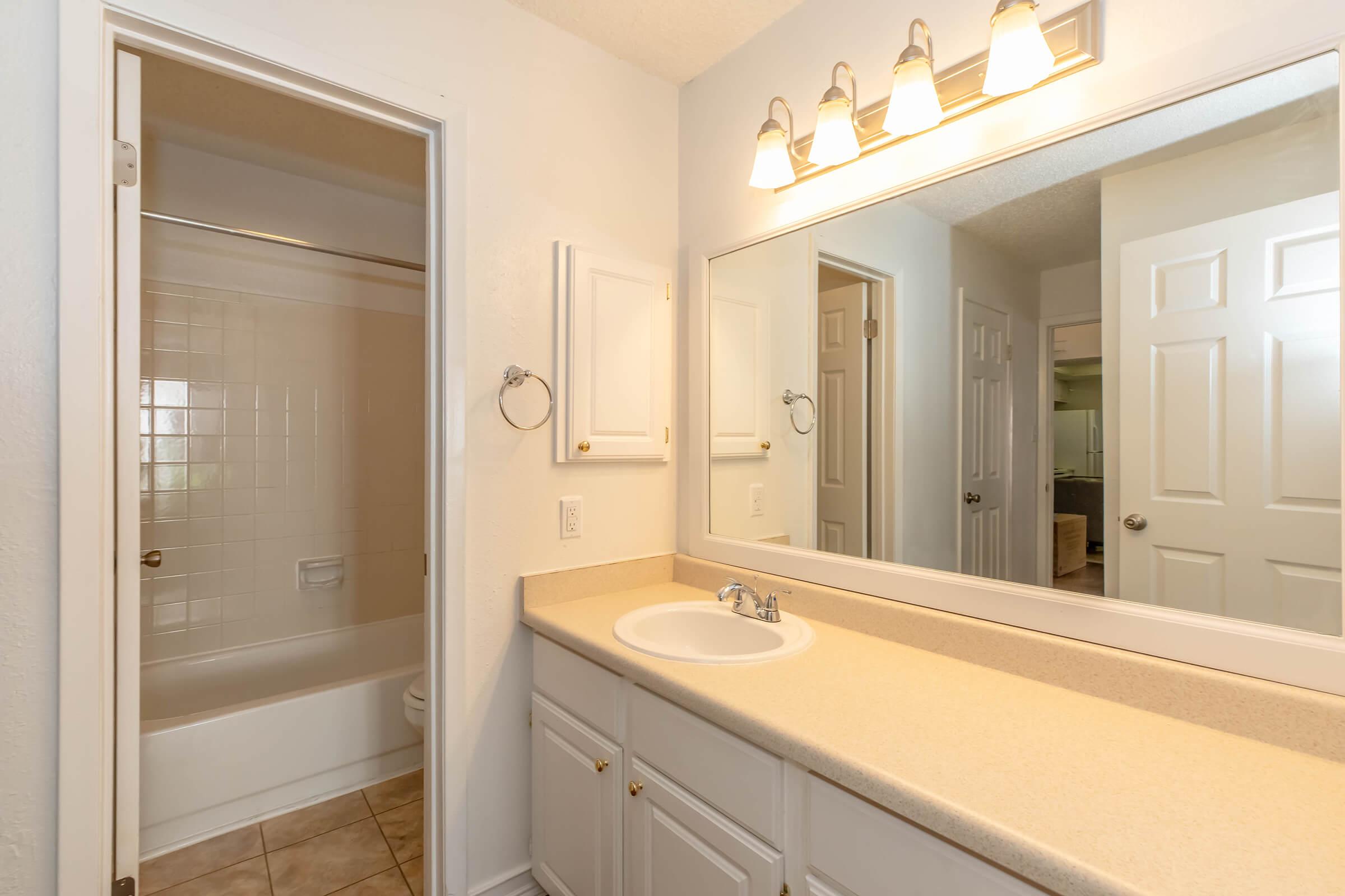 A well-lit bathroom featuring a beige countertop with a sink, a large mirror above, and cabinets. To the left, there's a door leading to a bathtub and shower, while the opposite wall displays a doorway leading to another room. The walls are painted in a light color, contributing to a spacious feel.
