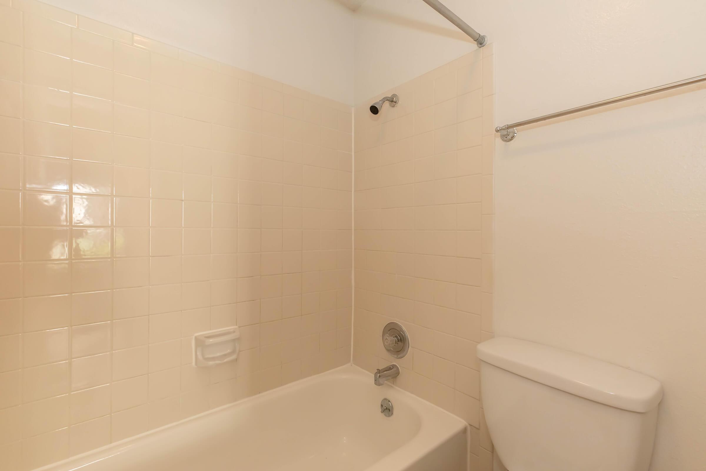 A simple bathroom with a beige tiled shower area and a bathtub underneath. There's a shower rod with a curtain, a faucet, and a toilet next to the bathtub. The walls are light-colored, creating a clean and minimalistic atmosphere.