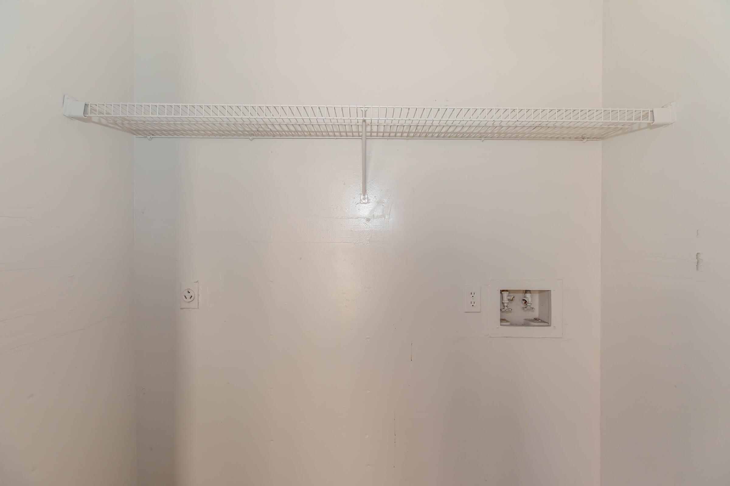 A blank room with white walls featuring a wire shelf mounted on the wall. There are two electrical outlet covers visible below the shelf, and a small cutout in the wall, possibly for plumbing or a utility connection. The overall appearance is minimal and uncluttered.