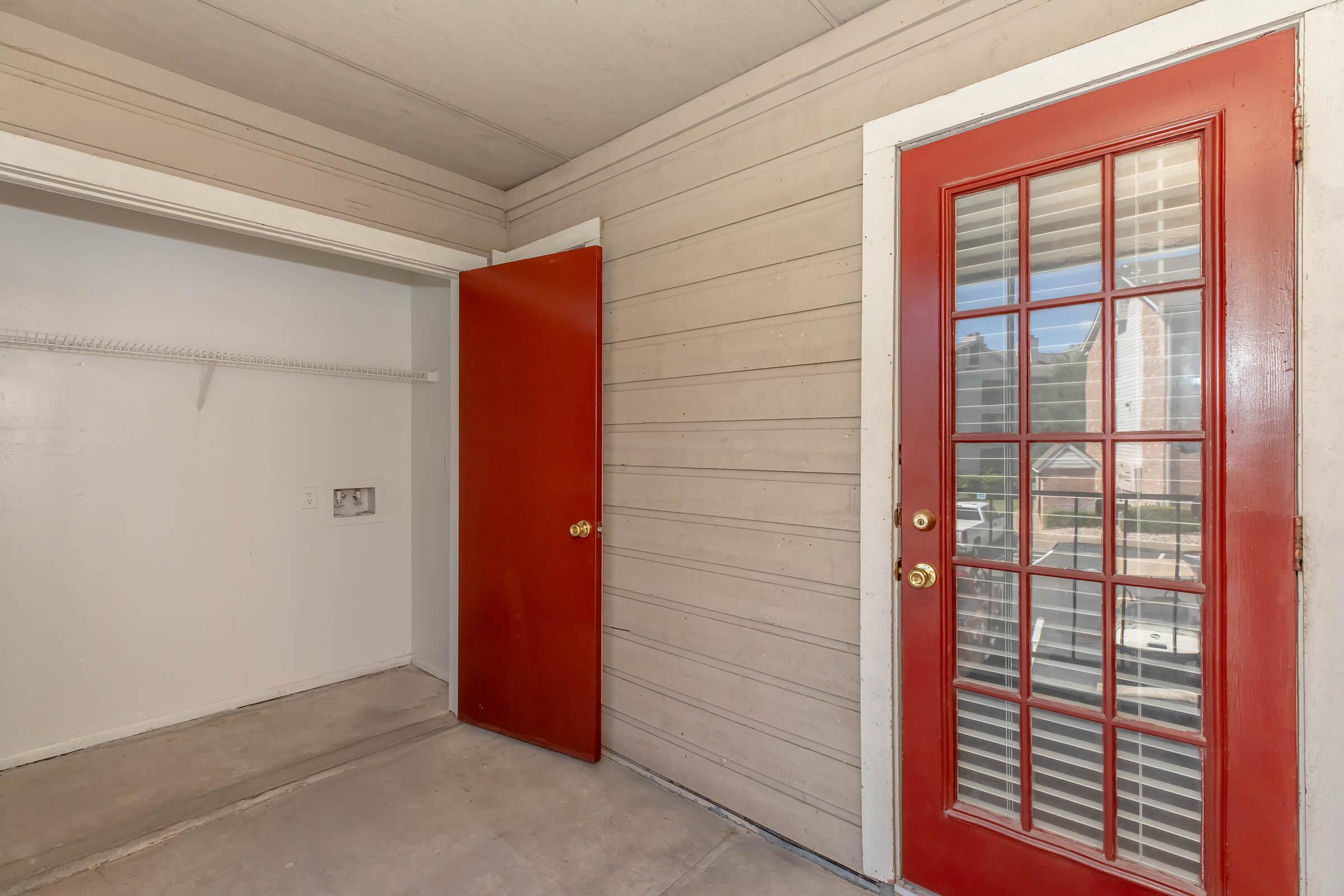 Interior view of a small room with light-colored wooden walls. One side features a closet with an open door, while the opposite side has a door with red frame and decorative glass panels leading outside. The floor is bare concrete, indicating the space may be in the process of renovation or preparation for new furnishings.