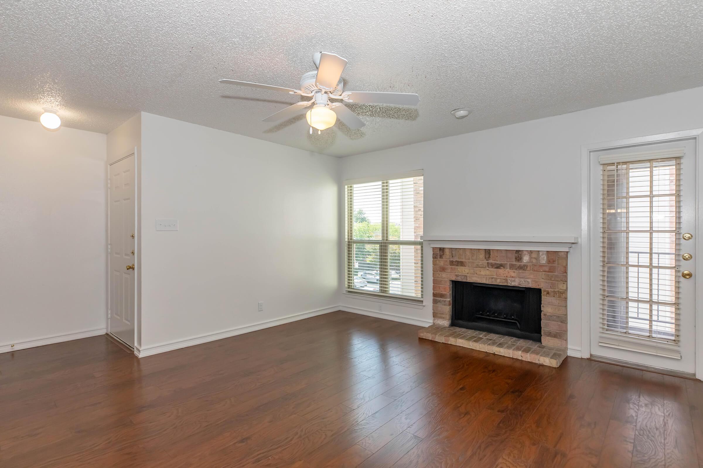 A spacious living area featuring hardwood floors, a ceiling fan, and a cozy brick fireplace. Large windows allow natural light to brighten the room. There are two doors: one leading to another room and another providing access to the exterior. The walls are painted white, creating a clean and inviting atmosphere.