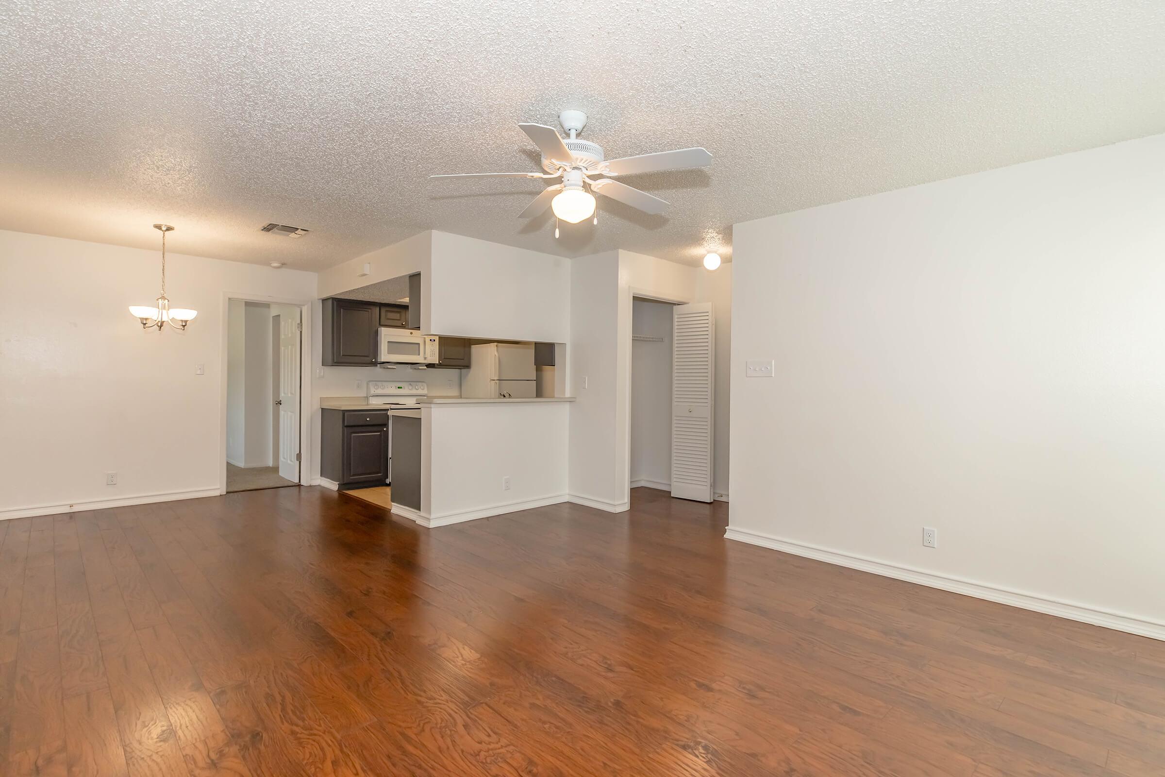A spacious living area featuring hardwood floors, a ceiling fan, and an open layout connecting to a kitchen with dark cabinets and white appliances. The room is brightly lit, with a dining area visible in the background. Walls are painted in a light color, enhancing the airy feel of the space.