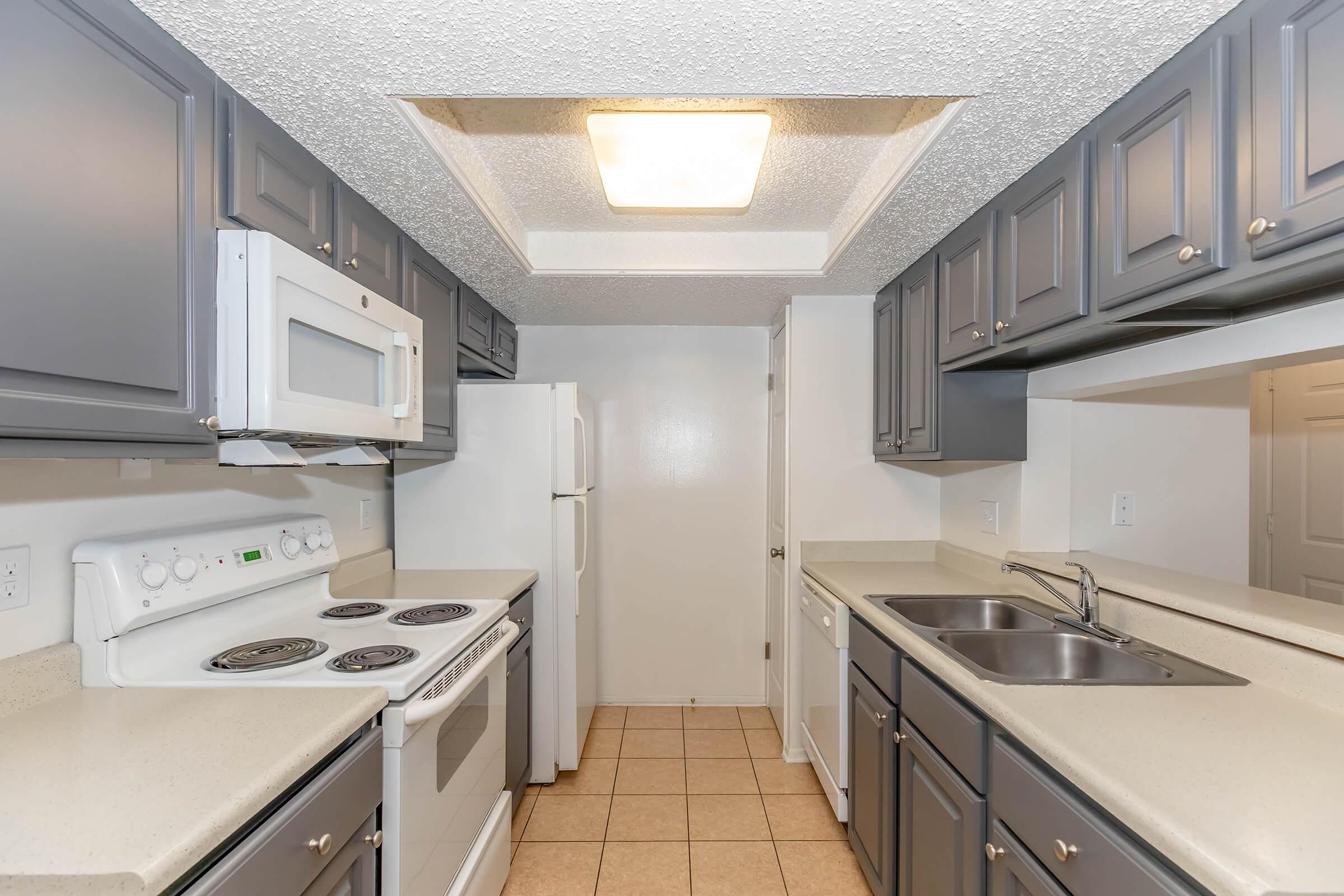 A modern kitchen featuring dark grey cabinetry, a white electric stove, microwave, and refrigerator. The countertops are light-colored, and the tile flooring is beige. The ceiling has a textured finish with a central light fixture, creating a bright and functional cooking space.