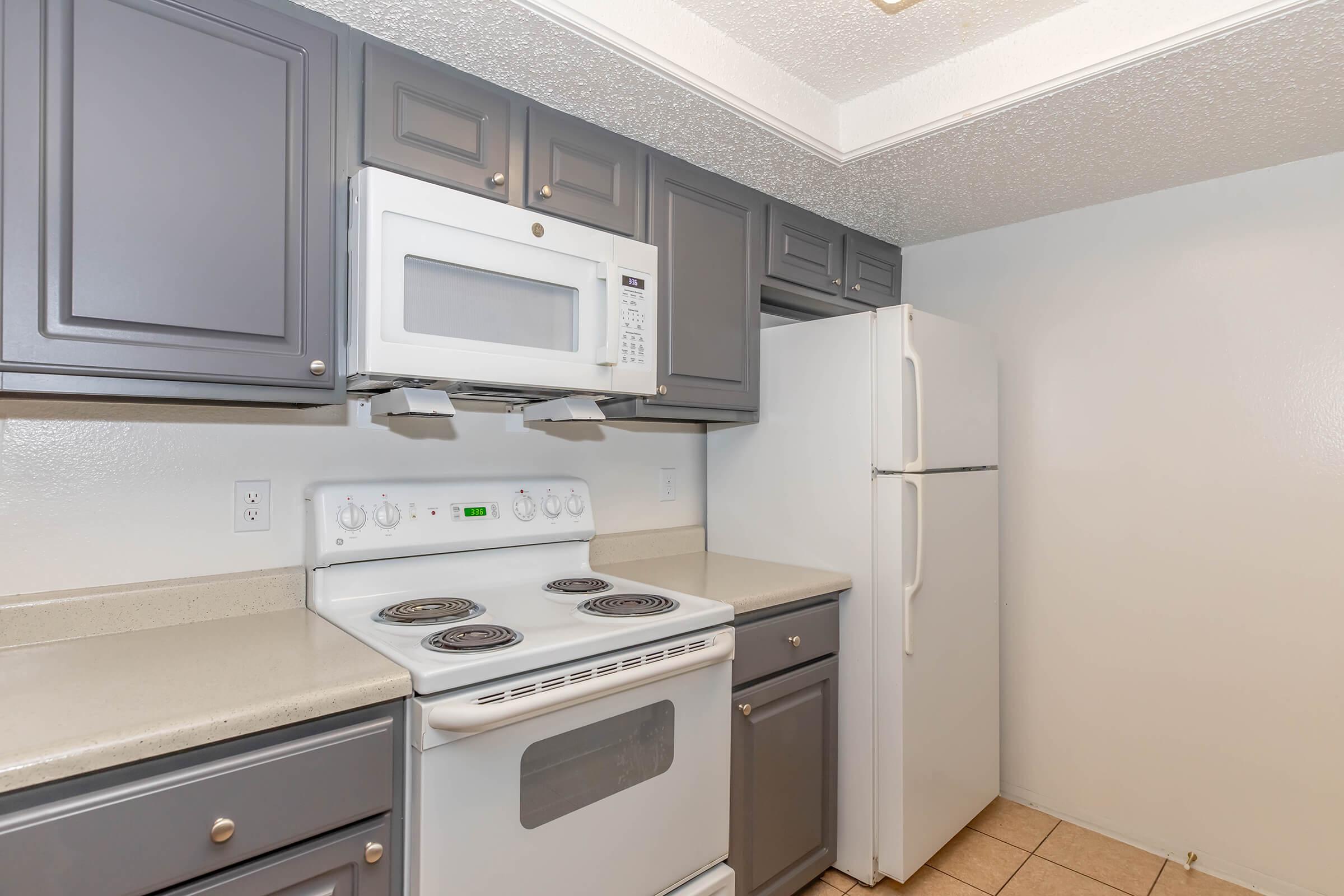 A modern kitchen featuring gray cabinets, a white stove with a microwave above, and a white refrigerator. The countertops are light-colored, and the flooring consists of beige tiles, creating a clean and functional cooking space. The walls are painted a light color, enhancing the brightness of the kitchen.