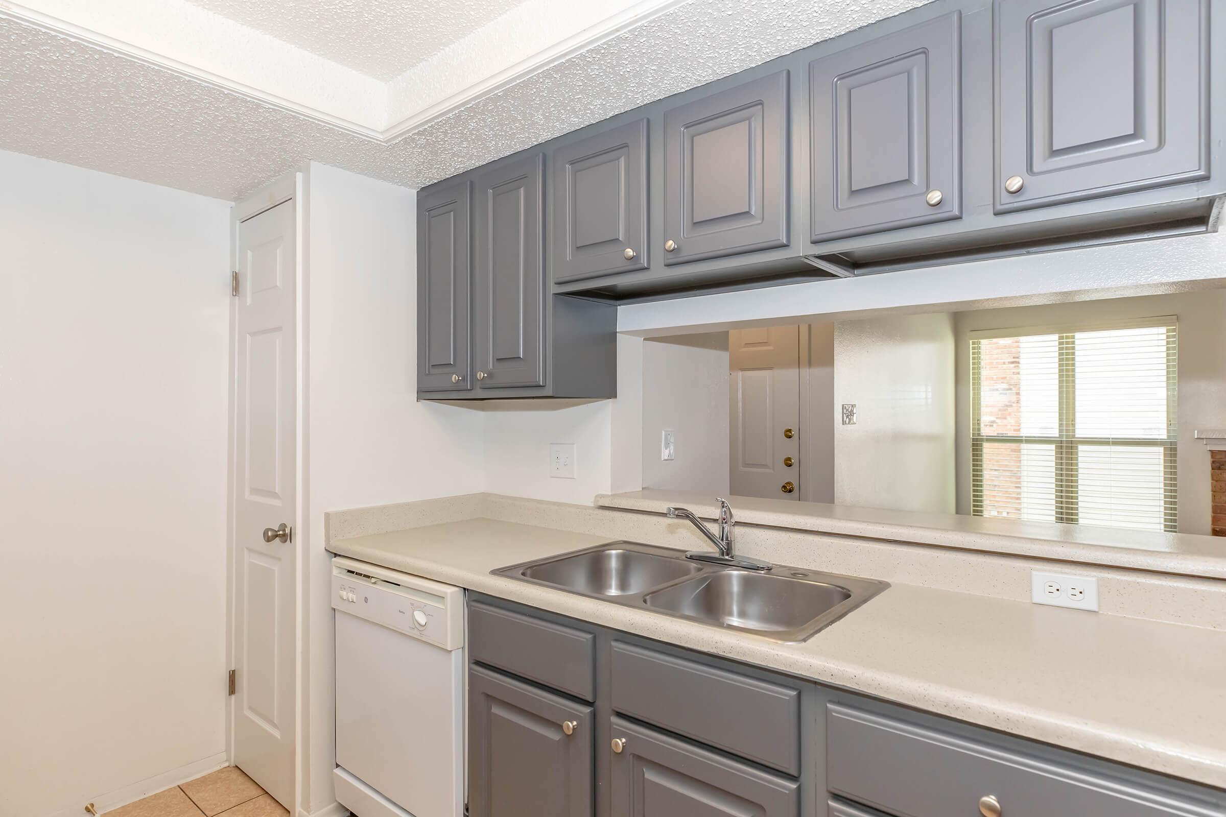 A modern kitchen featuring dark gray cabinets with silver knobs, a beige countertop, a double sink, and a dishwasher. The wall behind the cabinets is painted white, and there is a glimpse of a living space with large windows in the background. The flooring is light-colored tile.