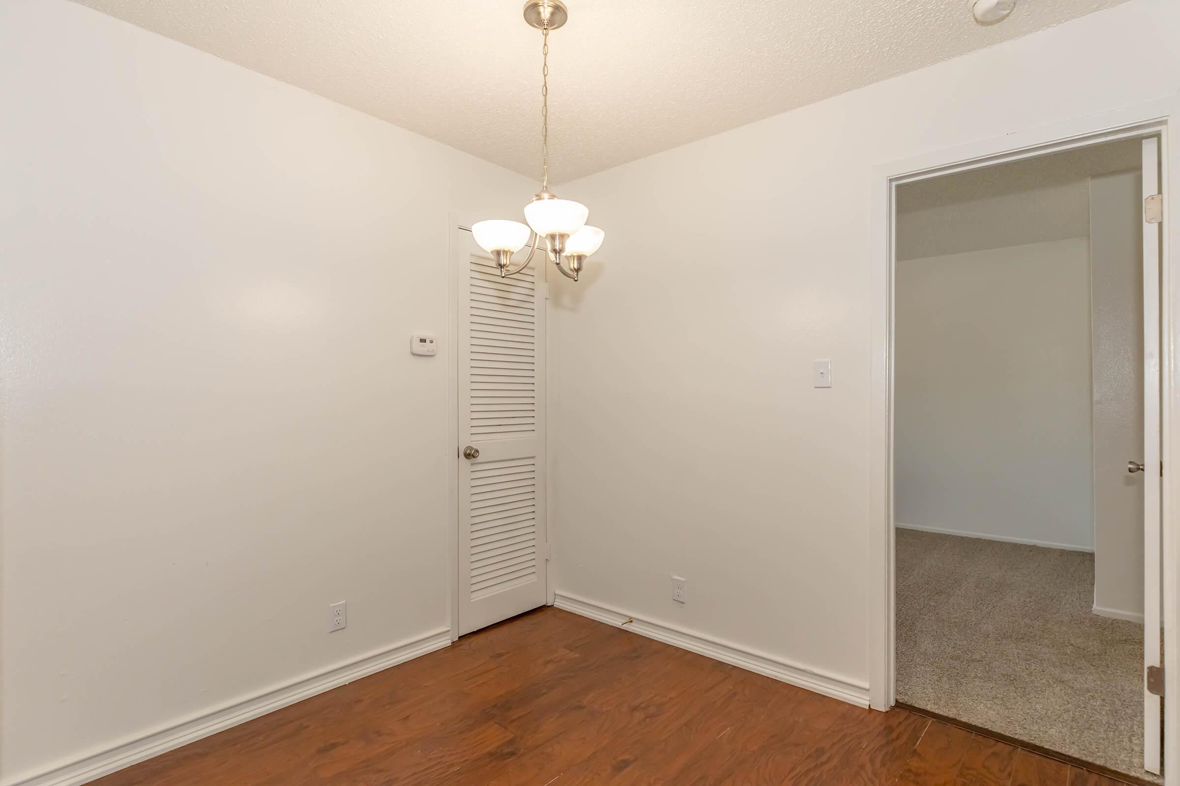 A simple interior space featuring hardwood flooring, a light fixture with three bulbs hanging from the ceiling, and a partially open door leading to another room. The walls are painted white, and there is a small, louvered door on the left side. The area appears clean and well-lit.