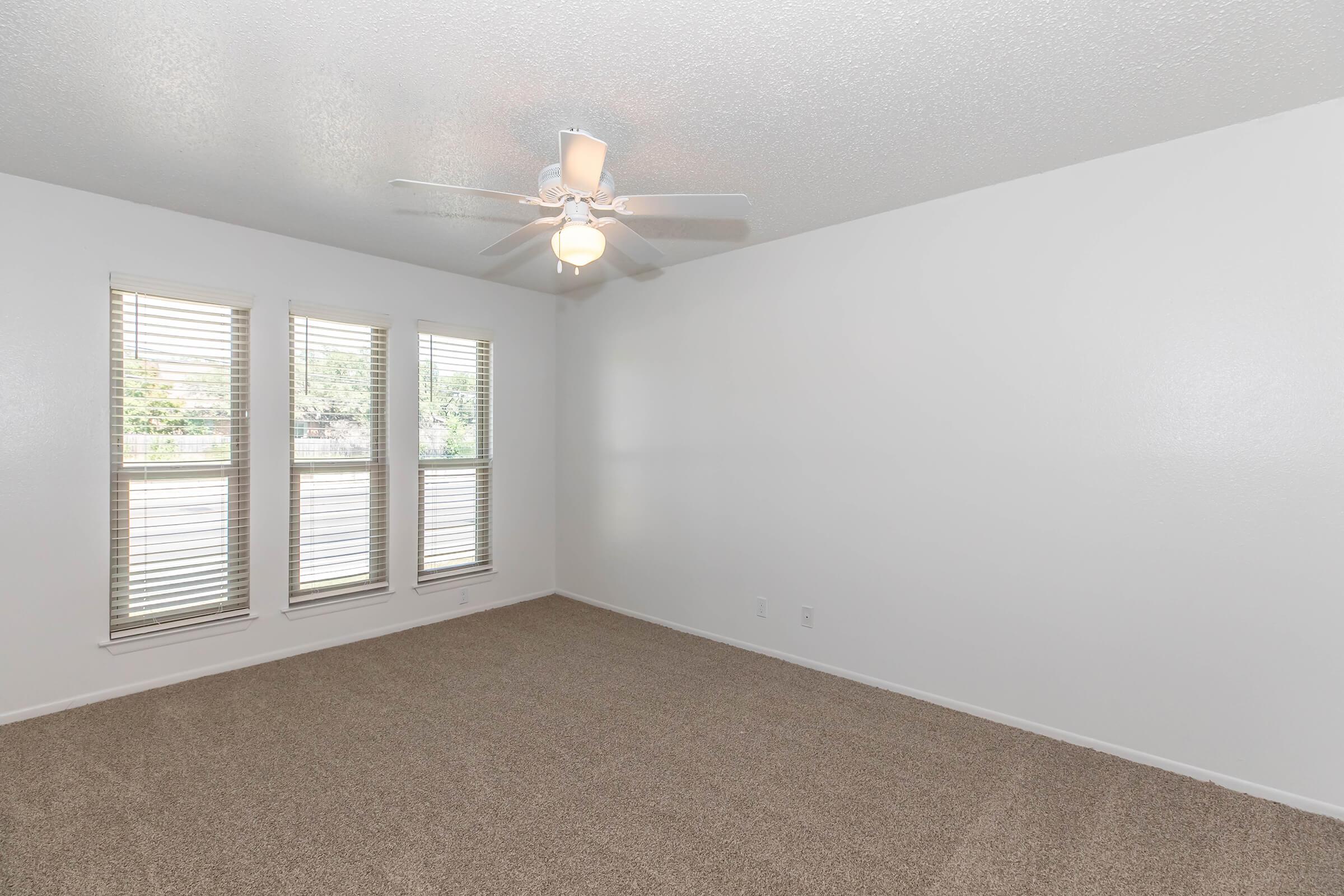 Interior view of a spacious, well-lit room with beige carpet flooring. It features three windows with blinds allowing natural light and a ceiling fan for ventilation. The walls are painted white, creating a bright and airy atmosphere, and the room is empty, providing a blank canvas for decoration.