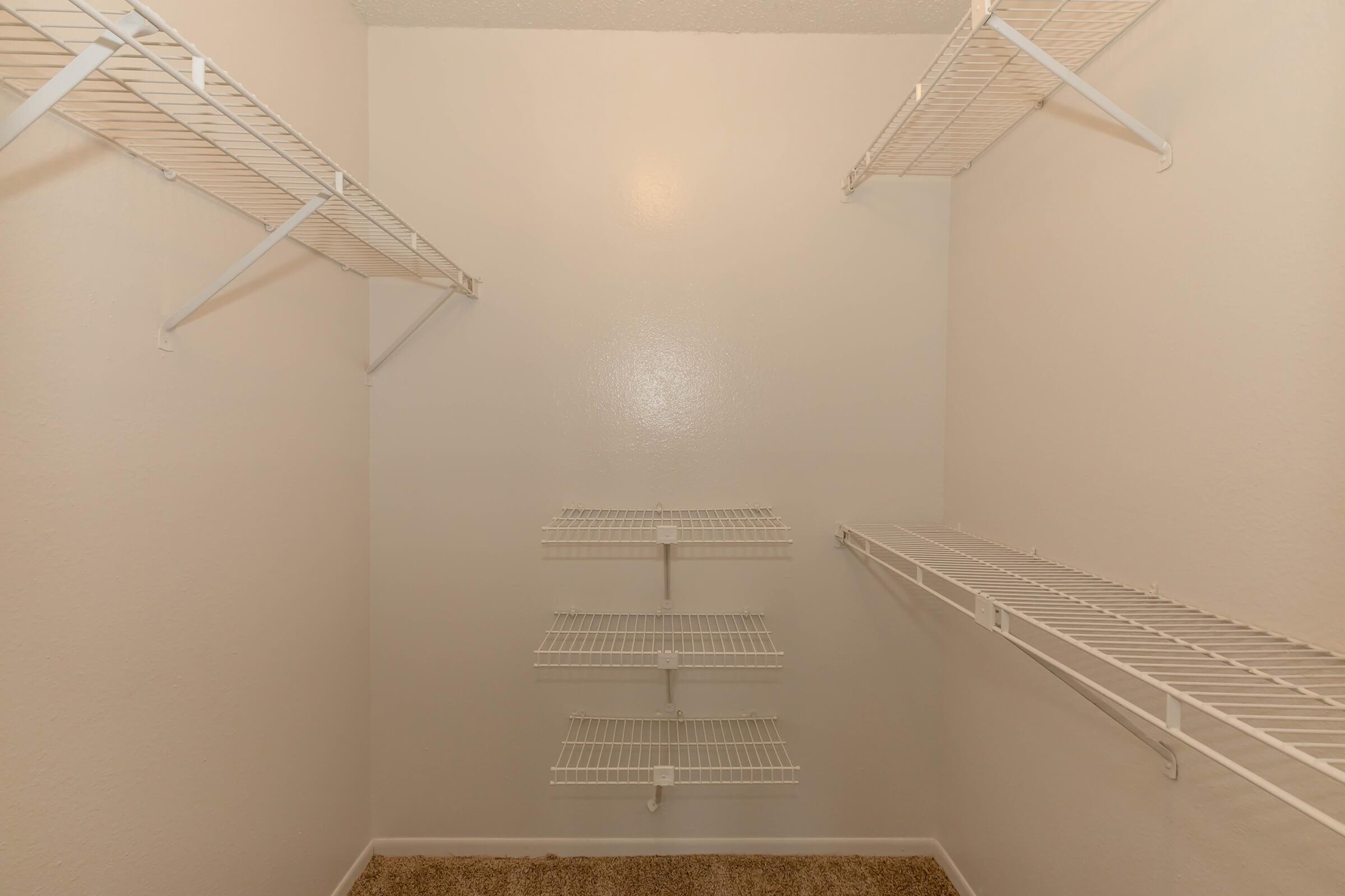 A clean, empty closet with white wire shelving mounted on the walls. The interior features multiple horizontal shelves on either side and a few vertical racks in the center. The walls are painted a light color, and the floor is covered with a beige carpet.