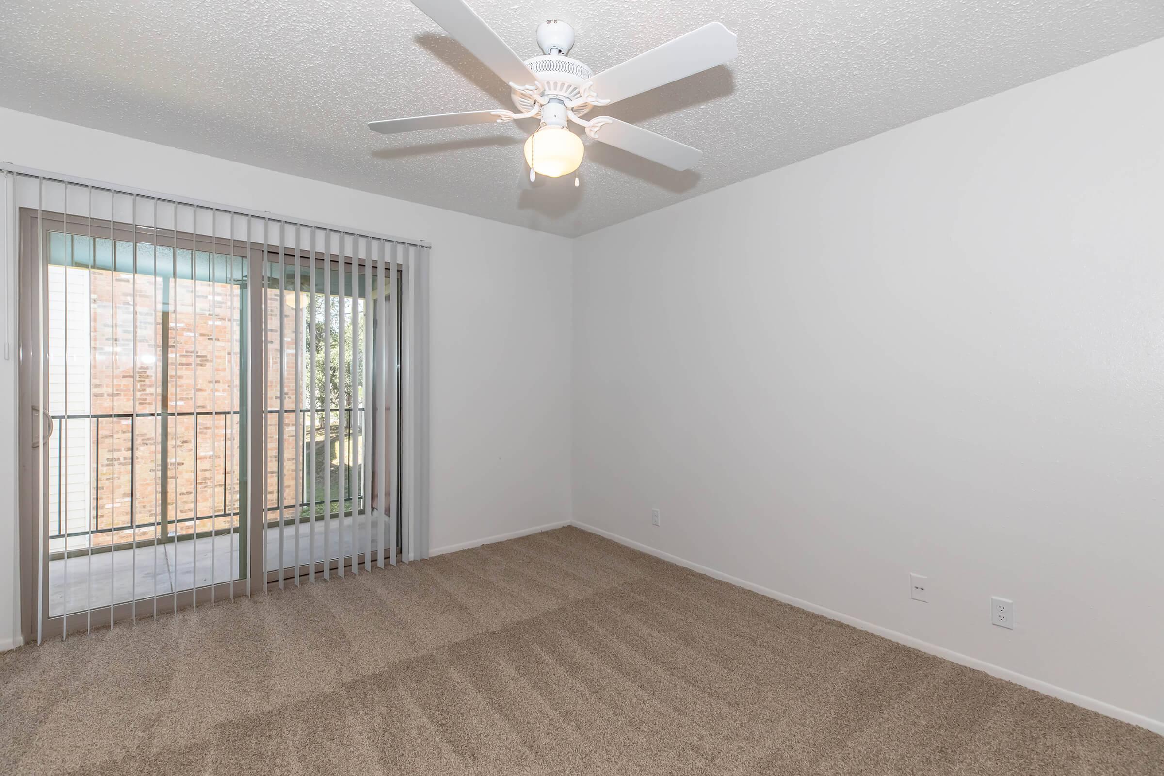 A clean, empty room with light beige carpet and white walls. A ceiling fan is installed in the center, and there is a sliding glass door with vertical blinds leading to a balcony. Natural light illuminates the space, enhancing the airy and open feel of the room.