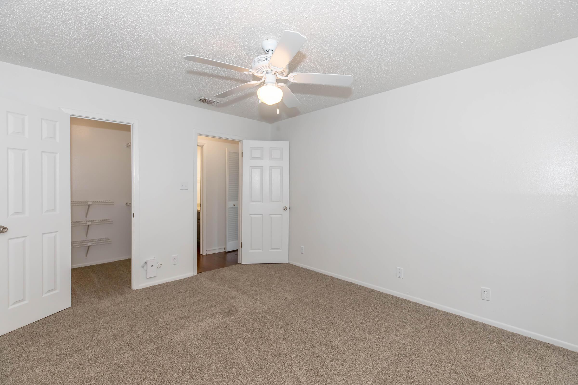 A clean and empty bedroom featuring light-colored walls, a ceiling fan, and beige carpet. There are two closed doors leading to closets or other rooms. Natural light illuminates the space, creating a bright and inviting atmosphere.