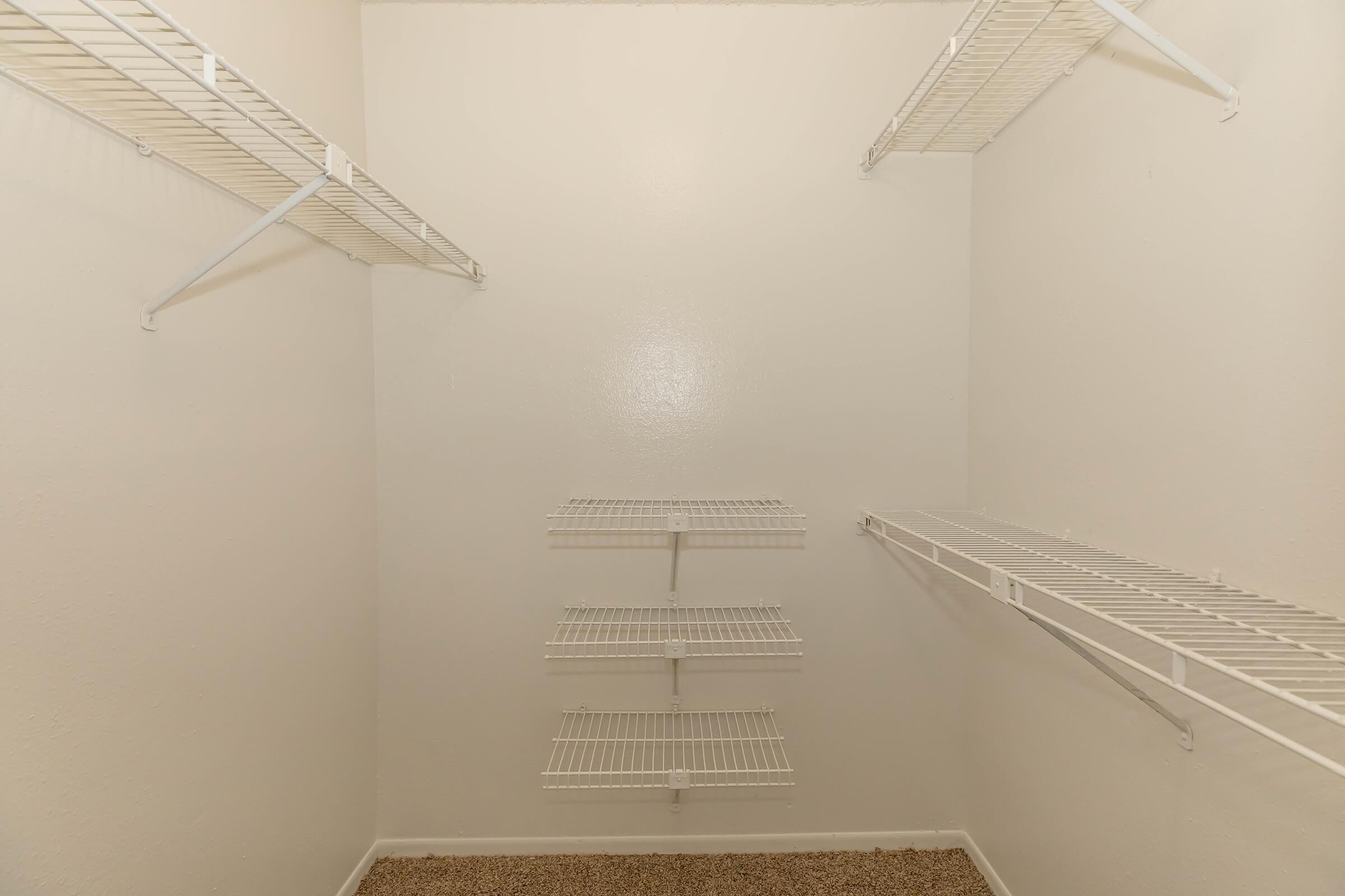 Empty closet with light-colored walls and tan carpet. There are several white wire shelves mounted on the walls, providing storage space. The closet appears clean and well-maintained, with no items inside.