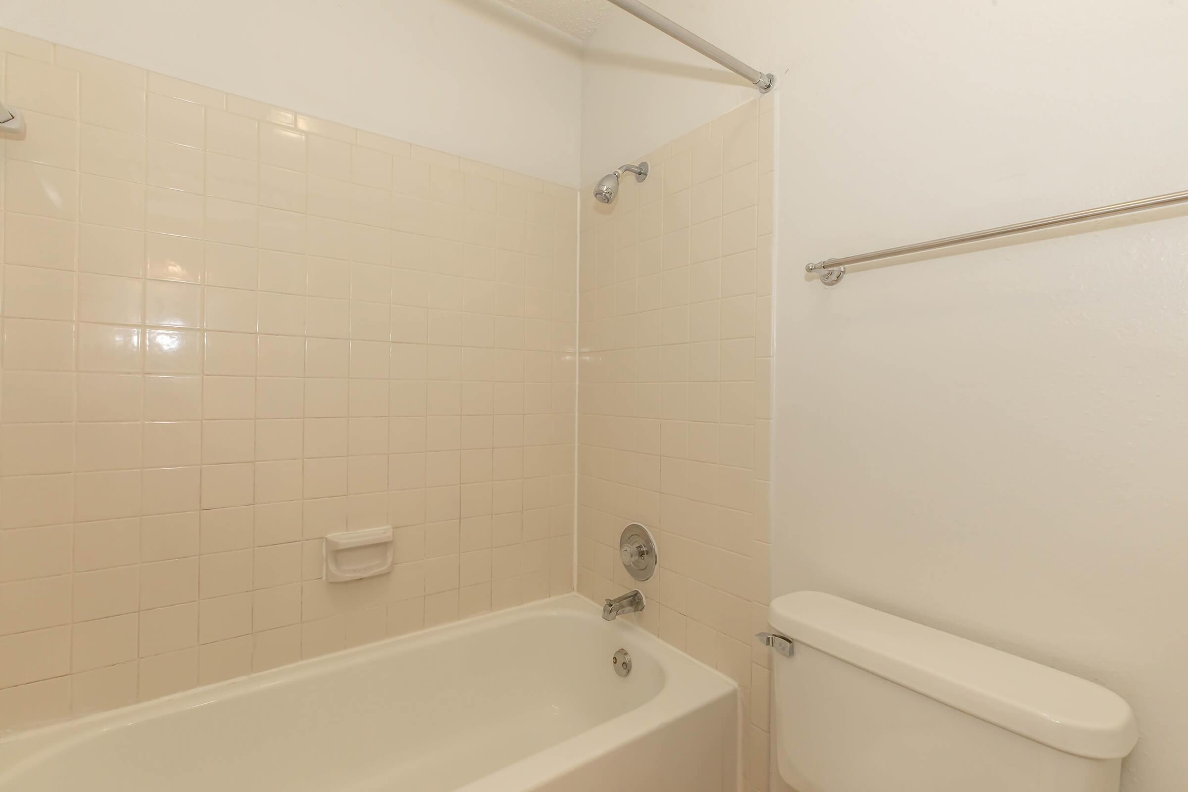 A clean, simple bathroom featuring a white bathtub with a showerhead above, light beige tiled walls, and a toilet nearby. A towel bar is mounted on the wall, and the overall decor is minimalist and functional. The space is well-lit with a neutral color palette.