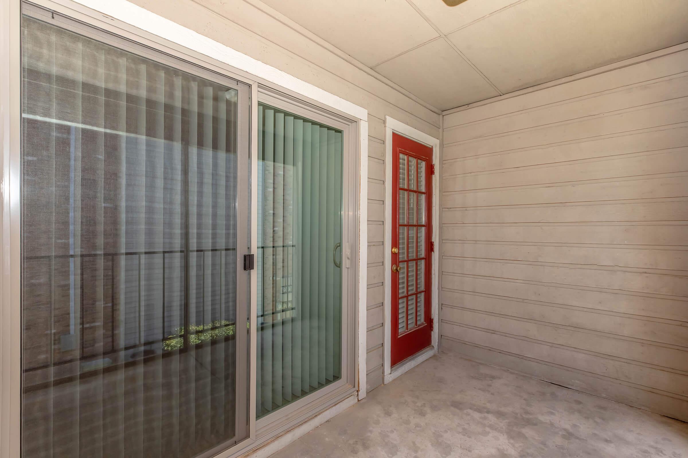 A covered outdoor patio area featuring sliding glass doors with vertical blinds, a red door with multiple panes, and textured beige walls. The floor is concrete, and there is natural light coming through the glass doors.