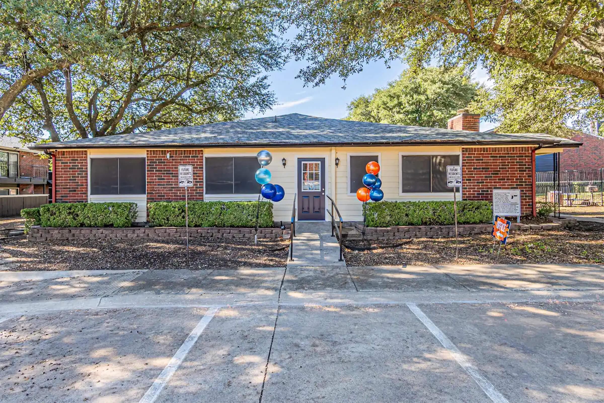 Single-story building with a brick facade, featuring large windows and a centered entrance. The front yard has neatly trimmed shrubs and two clusters of colorful balloons in blue, orange, and white. There are parking spaces in front, and signage visible on the property. Surrounding trees provide shade.