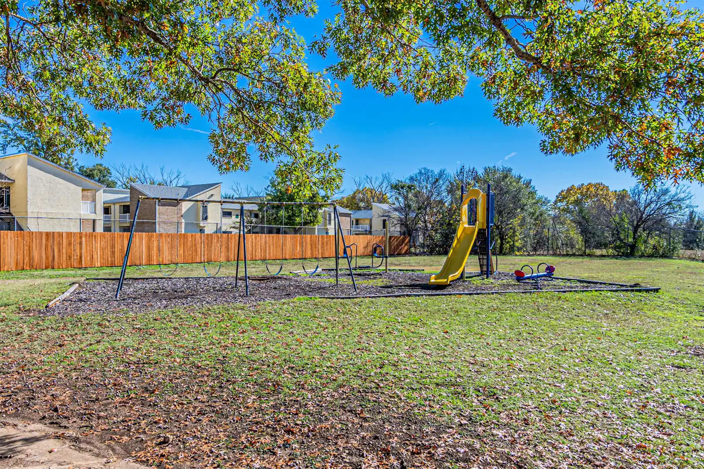 A sunny playground with a yellow slide and swings on a grassy area, surrounded by trees. In the background, there are apartment buildings and a wooden fence. The sky is clear and blue, creating a bright atmosphere.