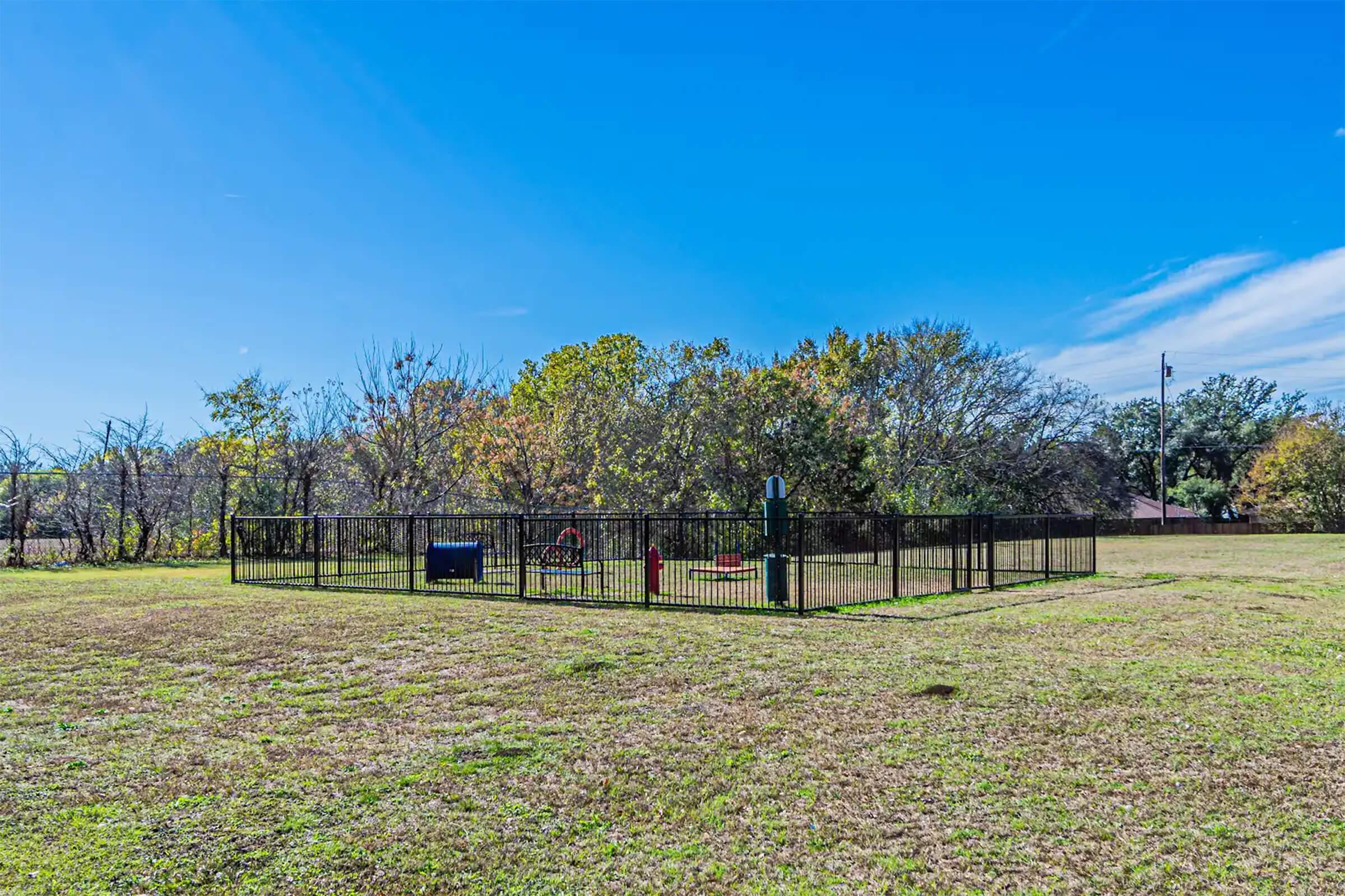 A fenced dog park located in a grassy area, surrounded by trees and under a clear blue sky. The park features play equipment for dogs, including agility structures, and there are some benches along the fence. The landscape is open with a few scattered trees in the background.