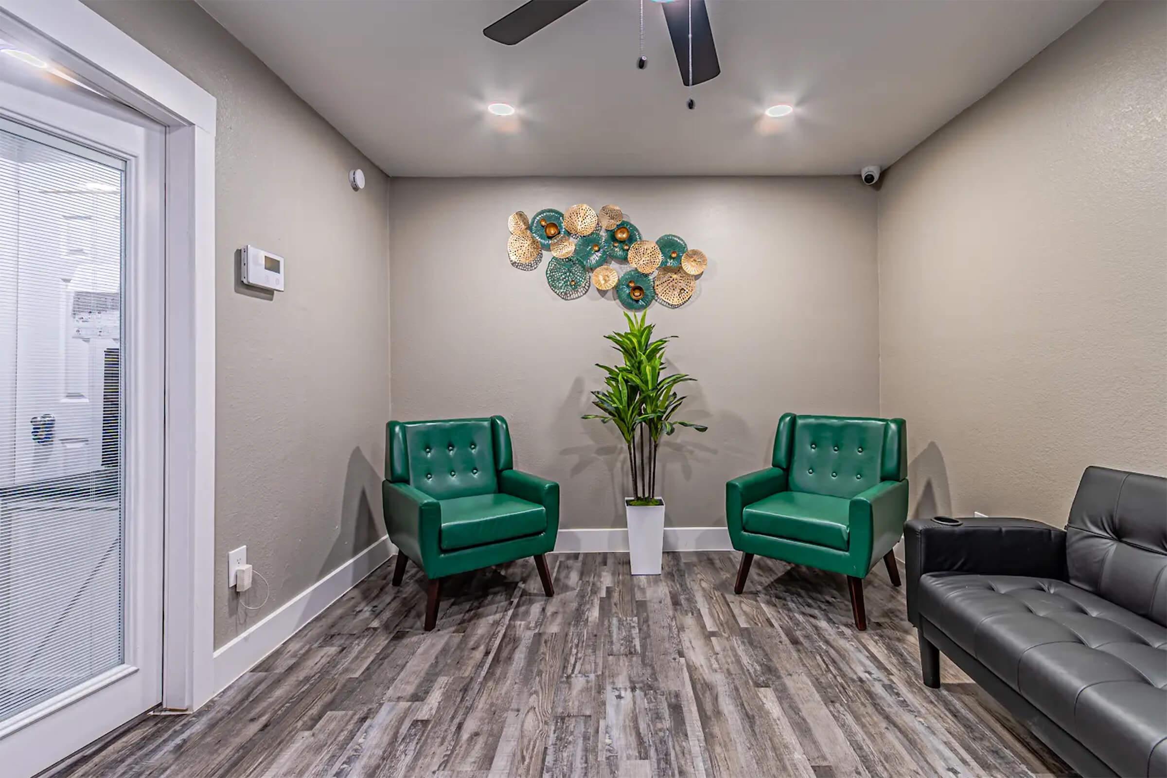 A cozy waiting area with two green upholstered chairs and a decorative wall art featuring circular shapes in shades of green and gold. A tall potted plant stands in the center, complementing the modern design. The flooring is a light brown wood-like material, enhancing the inviting atmosphere.