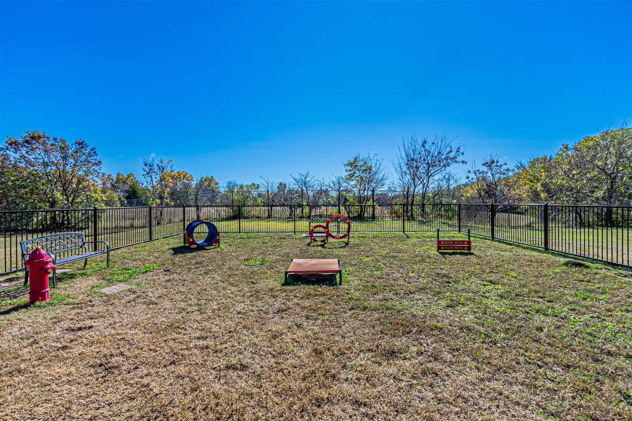 A grassy, fenced dog park featuring various play equipment, including a red fire hydrant, two colorful hoops, and a raised platform. Surrounding trees with autumn foliage are visible under a clear blue sky.