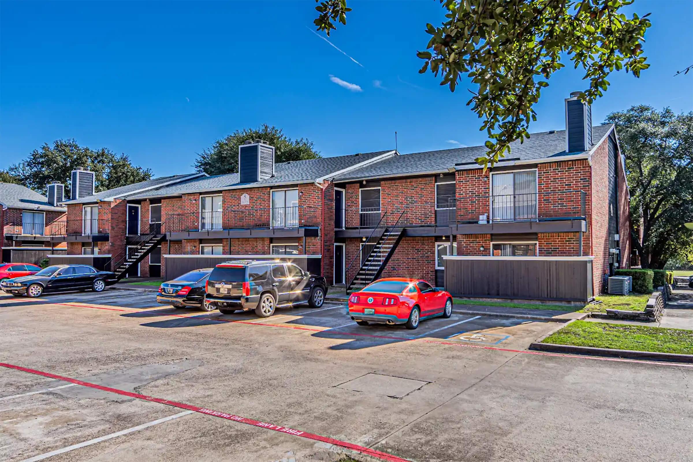 A well-maintained apartment complex featuring two-story brick buildings. The image shows several parked cars in front of the units, with balconies visible above. The surrounding area includes green grass and trees under a clear blue sky, indicating a sunny day.