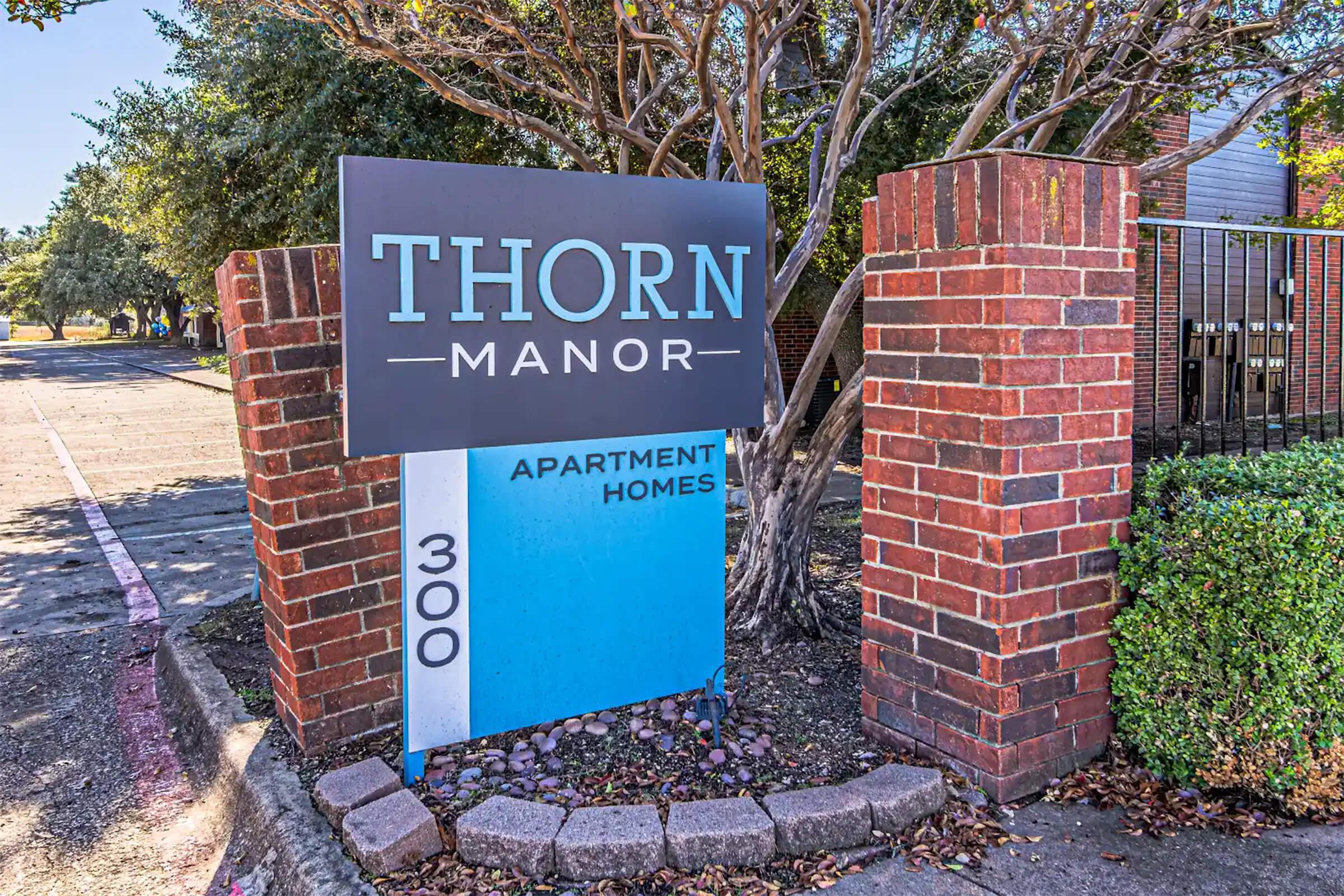Sign for "Thorn Manor" apartments, featuring a large blue and gray sign with the text "THORN MANOR" prominently displayed at the top and "APARTMENT HOMES" below it. The sign is surrounded by brick pillars and shrubbery, located at the entrance of the property.