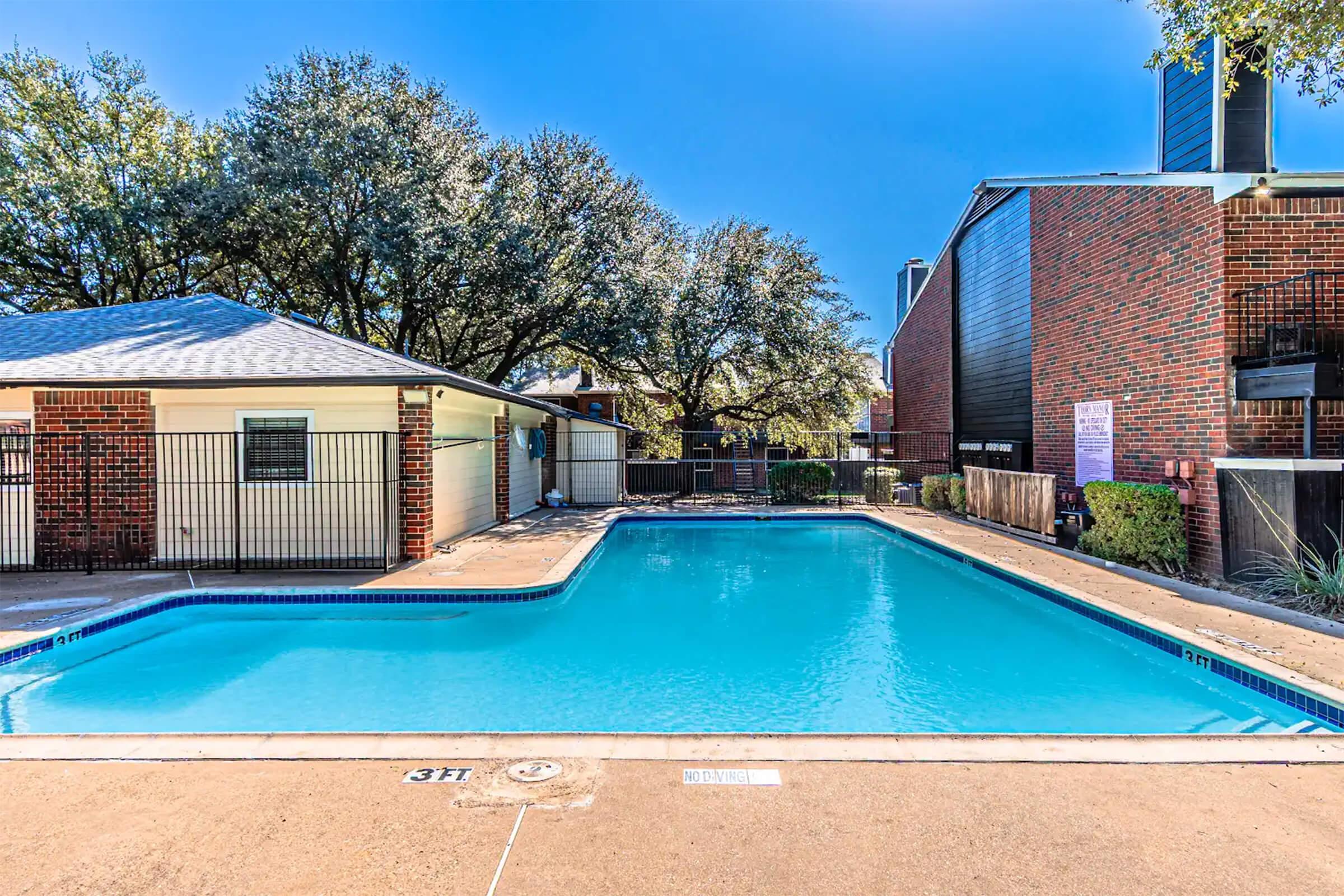 A clear blue swimming pool surrounded by a pool deck, residential buildings, and trees. The pool features a shallow end marked with a depth of 3 feet and has a clean, inviting appearance under a clear blue sky. Nearby structures include covered areas and a fence for privacy.