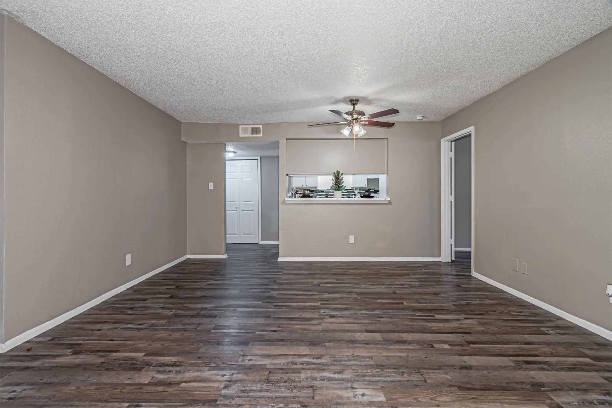 A spacious, empty living room with a textured ceiling and light brown walls. The floor is made of dark wood planks. A ceiling fan is installed in the center. In the background, there is a doorway leading to another room and an opening that looks into a kitchen area with a small plant on the counter.