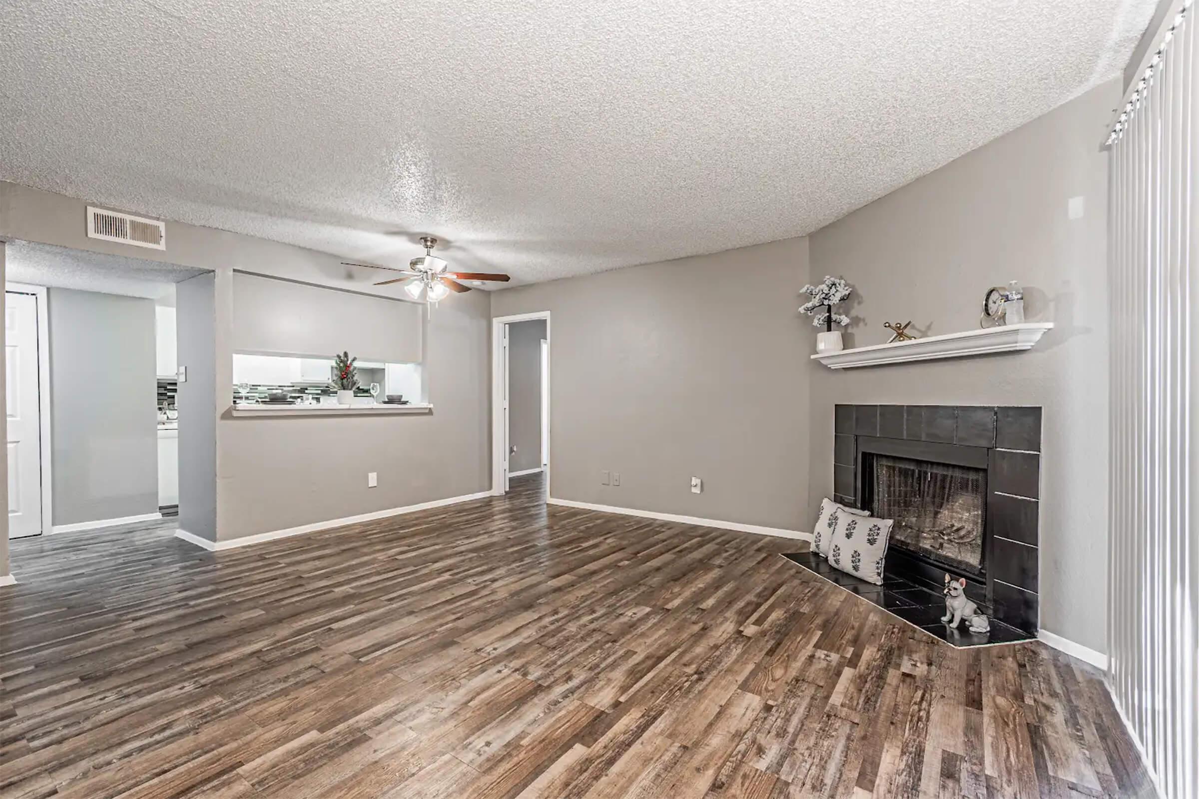 Spacious living room with a textured ceiling, light-colored walls, and wood laminate flooring. A black fireplace is situated on the right, adorned with decorative items. In the background, there's an open kitchen area visible. Natural light enters through a window on the left.