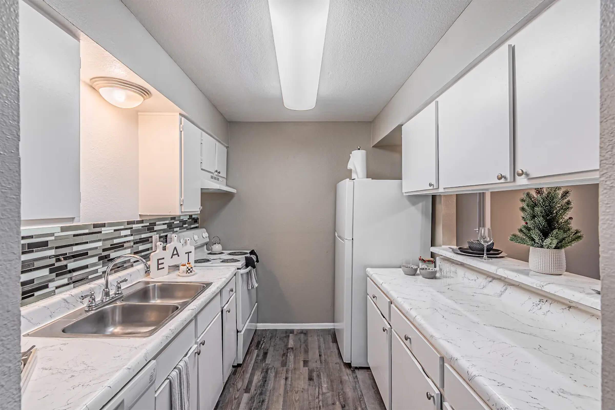 A modern kitchen featuring white cabinetry and a marble countertop. The space includes a double sink, a refrigerator, and an oven. A decorative backsplash adds a stylish touch, while a small plant and dinnerware are displayed on the counter. The walls are painted in a light color, enhancing the bright atmosphere.