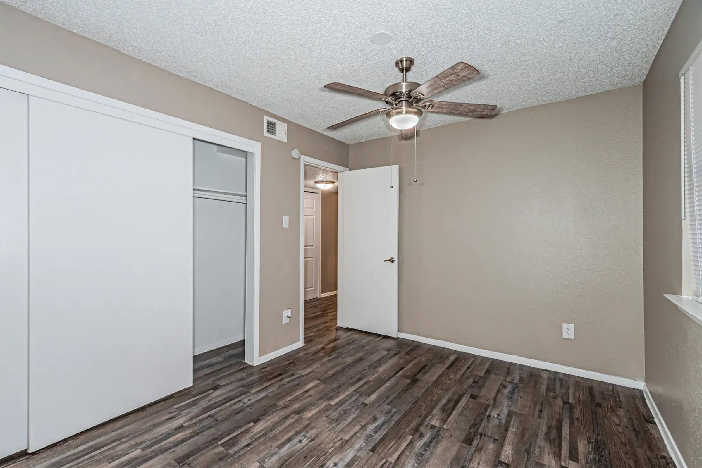 A small, empty room featuring a ceiling fan, light-colored walls, and laminate flooring. There is a closet with sliding doors on one side and a door leading to another room. Natural light enters through a window on the opposite wall. The space is well-lit and tidy, suitable for various uses.
