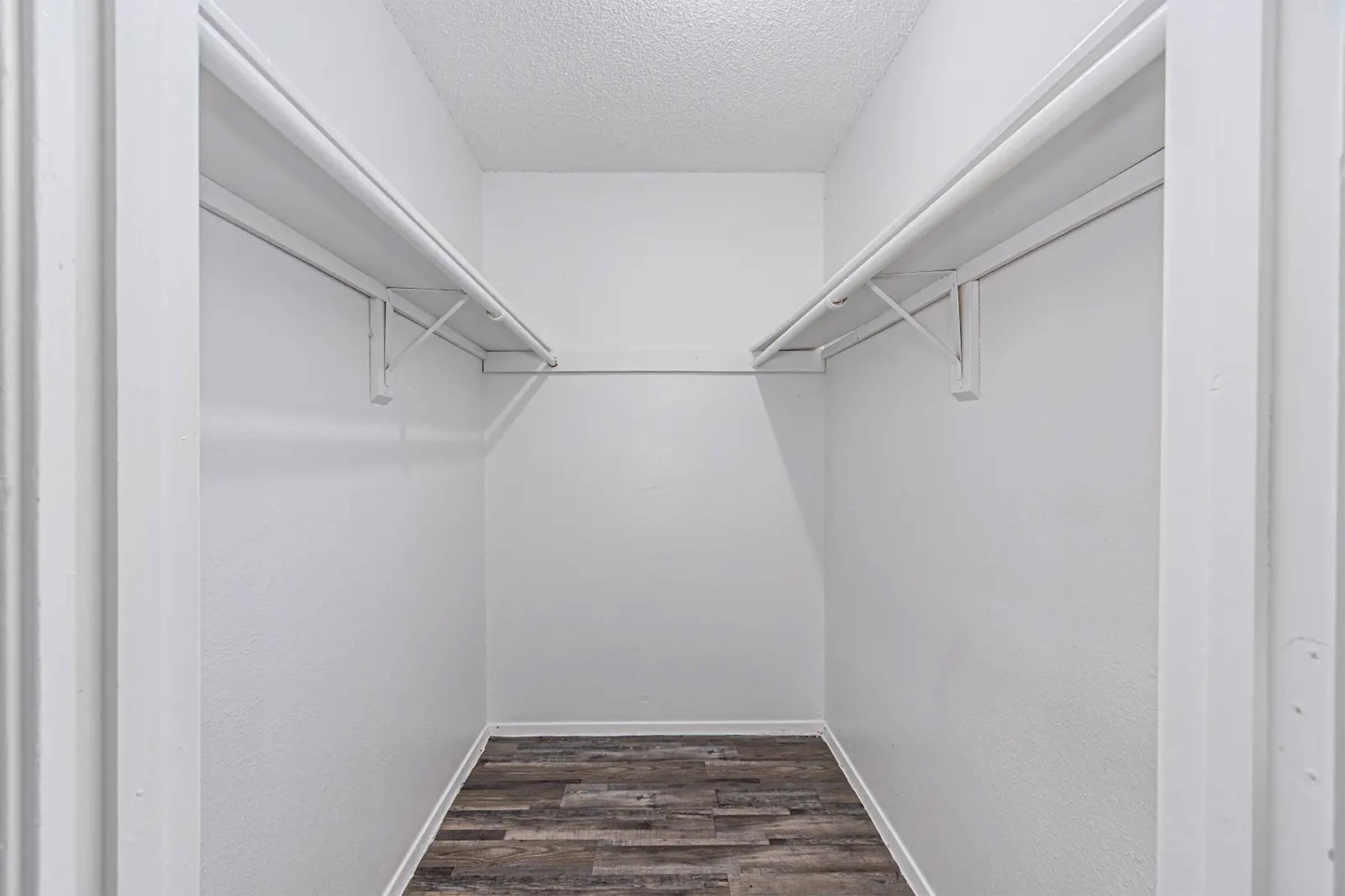 Empty walk-in closet with white walls and two hanging rods. The floor is covered with dark wood-like planks, creating a clean and spacious look. The overall atmosphere is bright and minimalistic, ideal for organizing clothing and accessories.