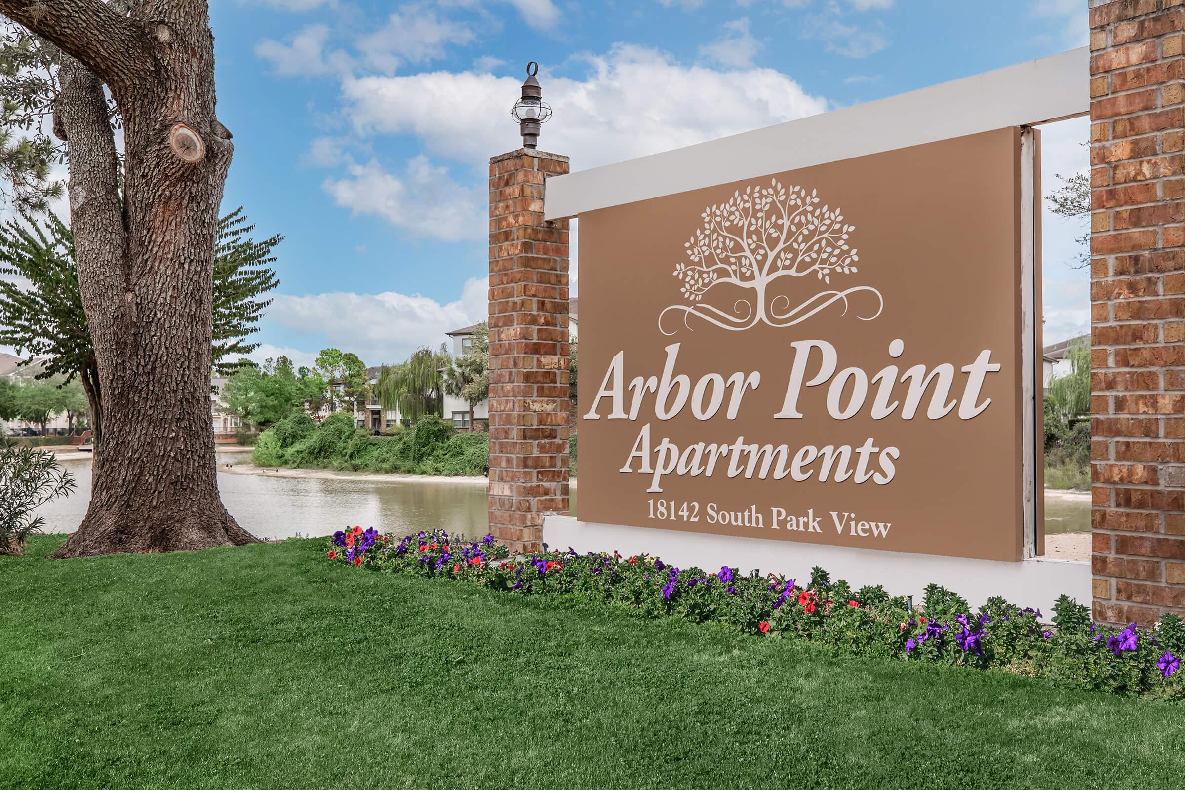 Sign for Arbor Point Apartments featuring the name prominently displayed, surrounded by landscaped greenery and colorful flowers. The background includes a tranquil water view and trees, creating a serene atmosphere.