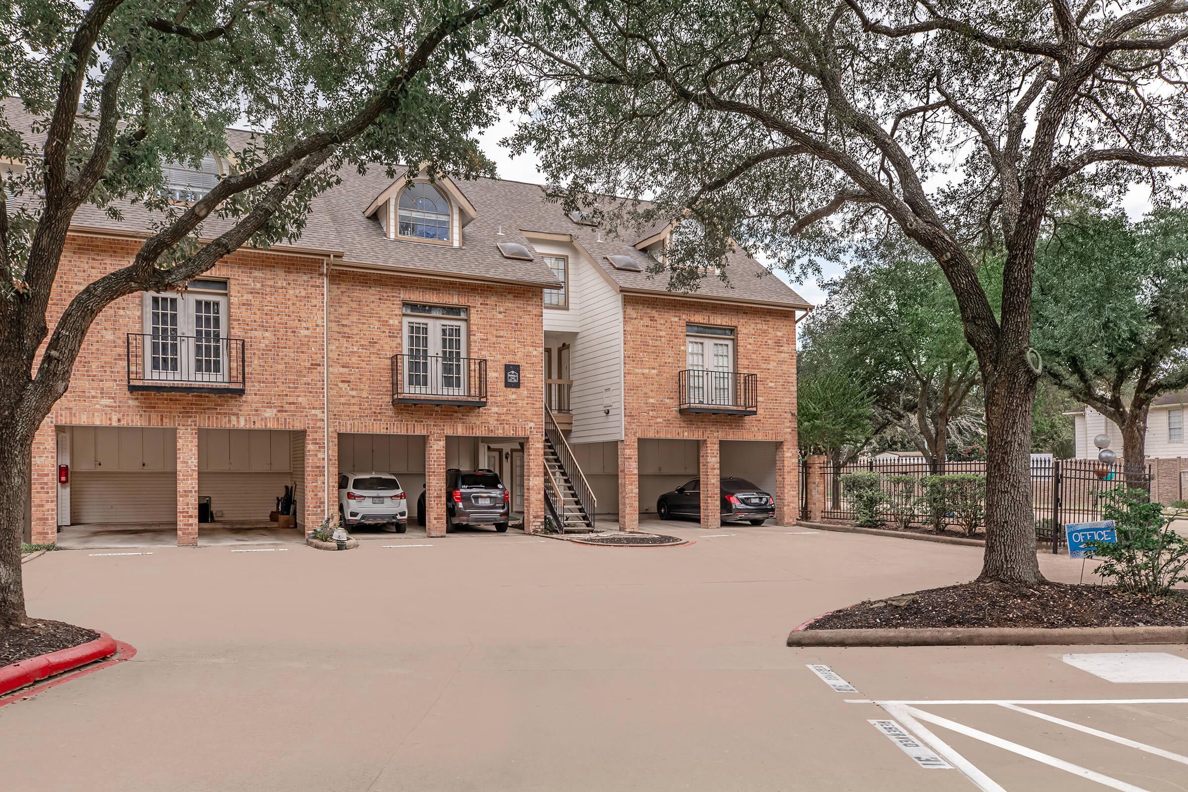 A view of a brick apartment complex with three units, each having private balconies. Below, carports shelter several parked vehicles. Surrounded by trees, the area is neatly landscaped, and a pool area is visible in the background. The parking lot is paved and features designated spaces.
