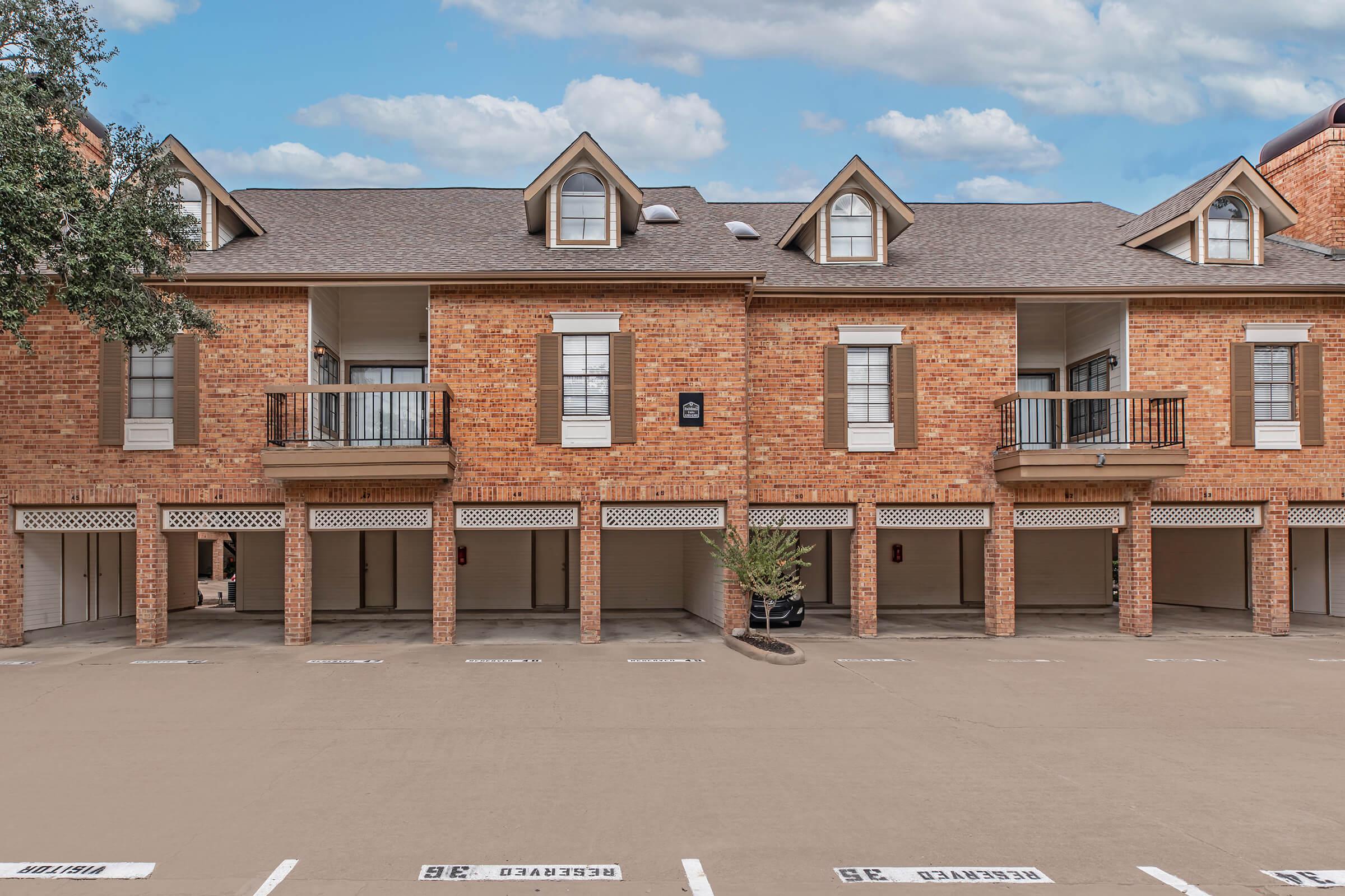 A row of brick townhouses with balconies and white shutters. The ground level features parking spaces, some marked with numbers. A small potted plant is visible near the entrance of one townhouse. The sky above is partly cloudy, creating a bright exterior setting.