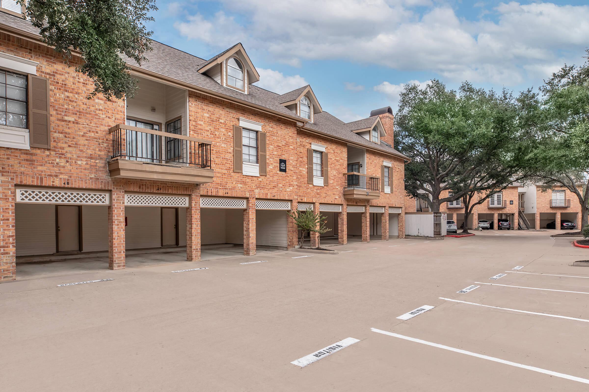 A row of brick apartment buildings with balconies, featuring garage spaces below. The parking area is lined with designated spaces, and there are trees nearby under a partly cloudy sky. The scene conveys a residential neighborhood atmosphere.